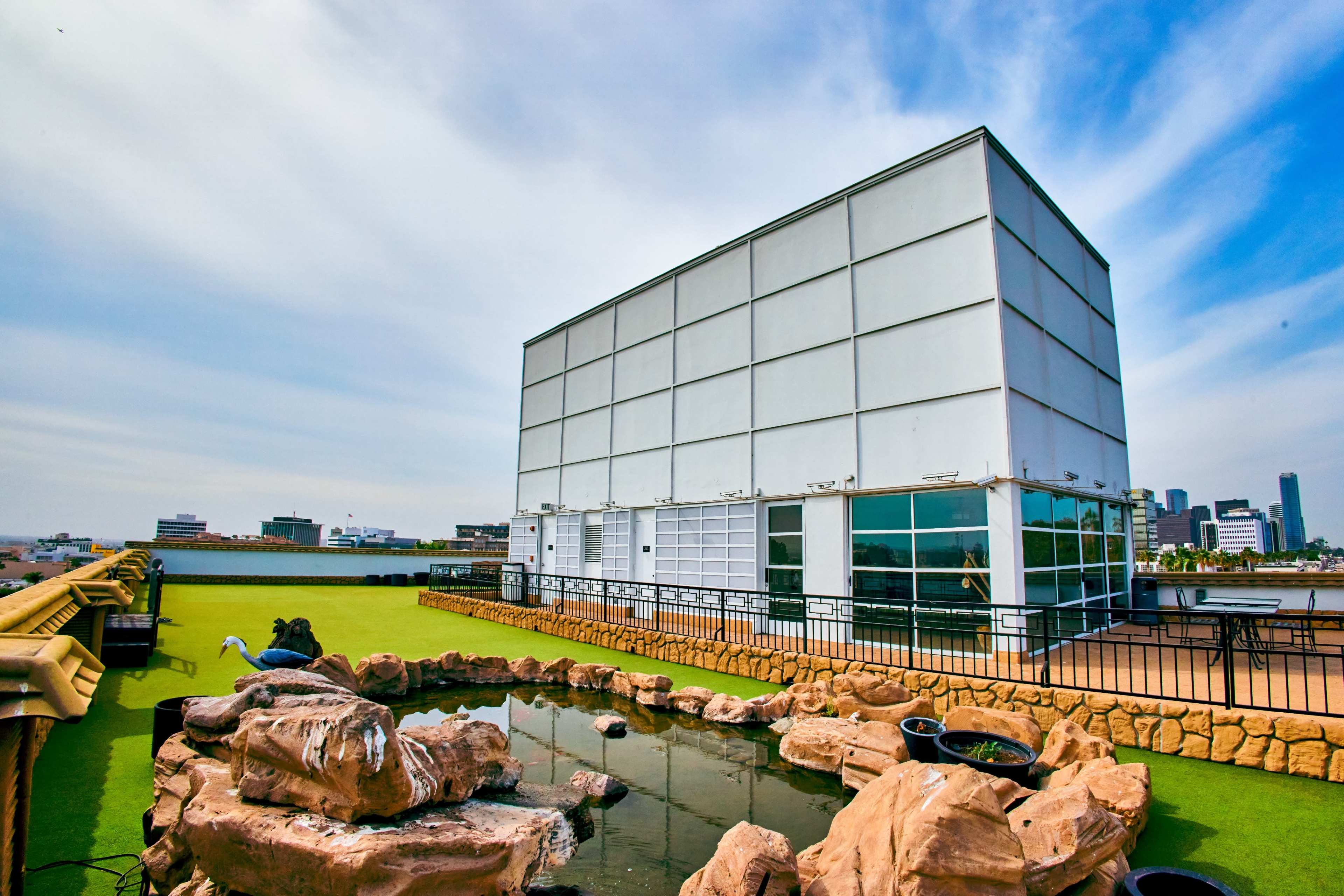 A modern white building with large windows is situated on a rooftop featuring a small, rocky pond and green landscaping.
