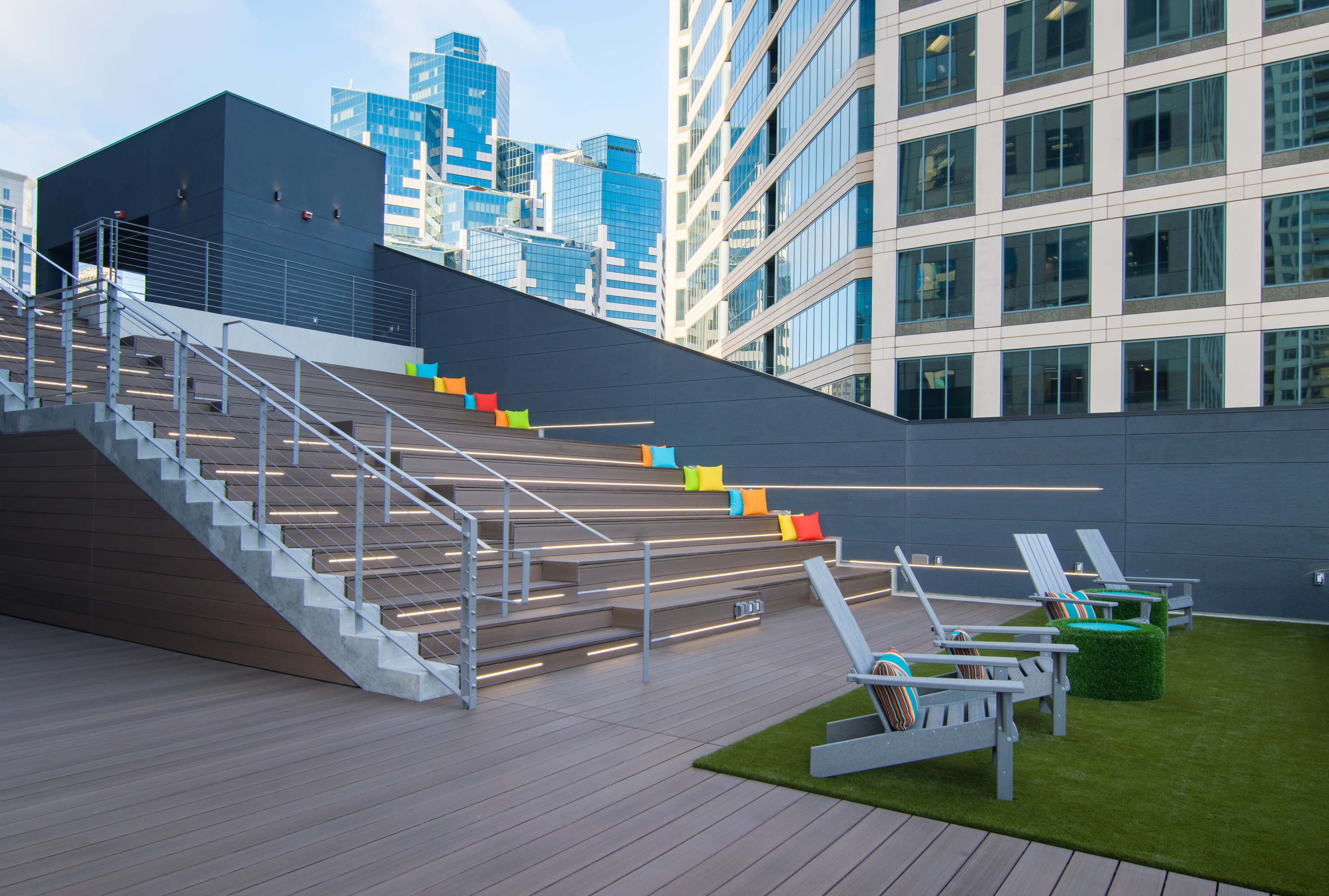 A modern rooftop area with colorful seating on the stairs, green turf, and lounge chairs amid a backdrop of tall buildings.