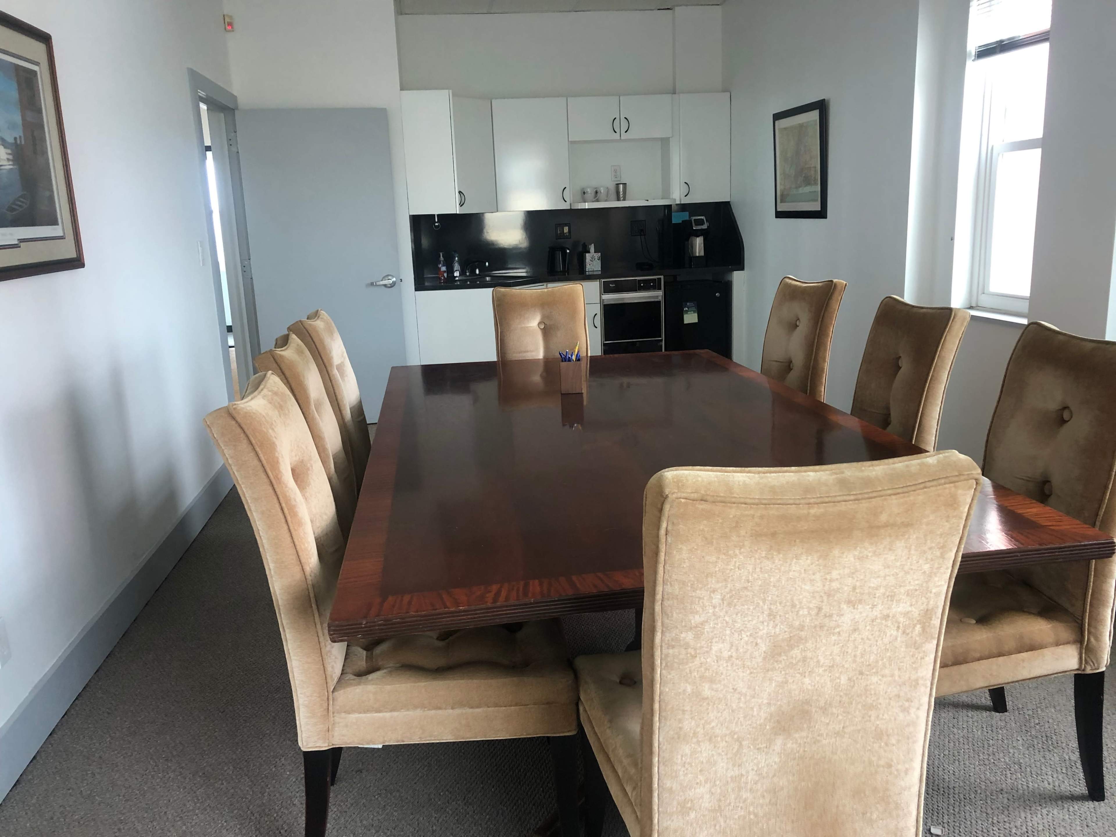 A large wooden conference table with eight upholstered chairs is situated in a well-lit meeting room featuring a kitchenette.