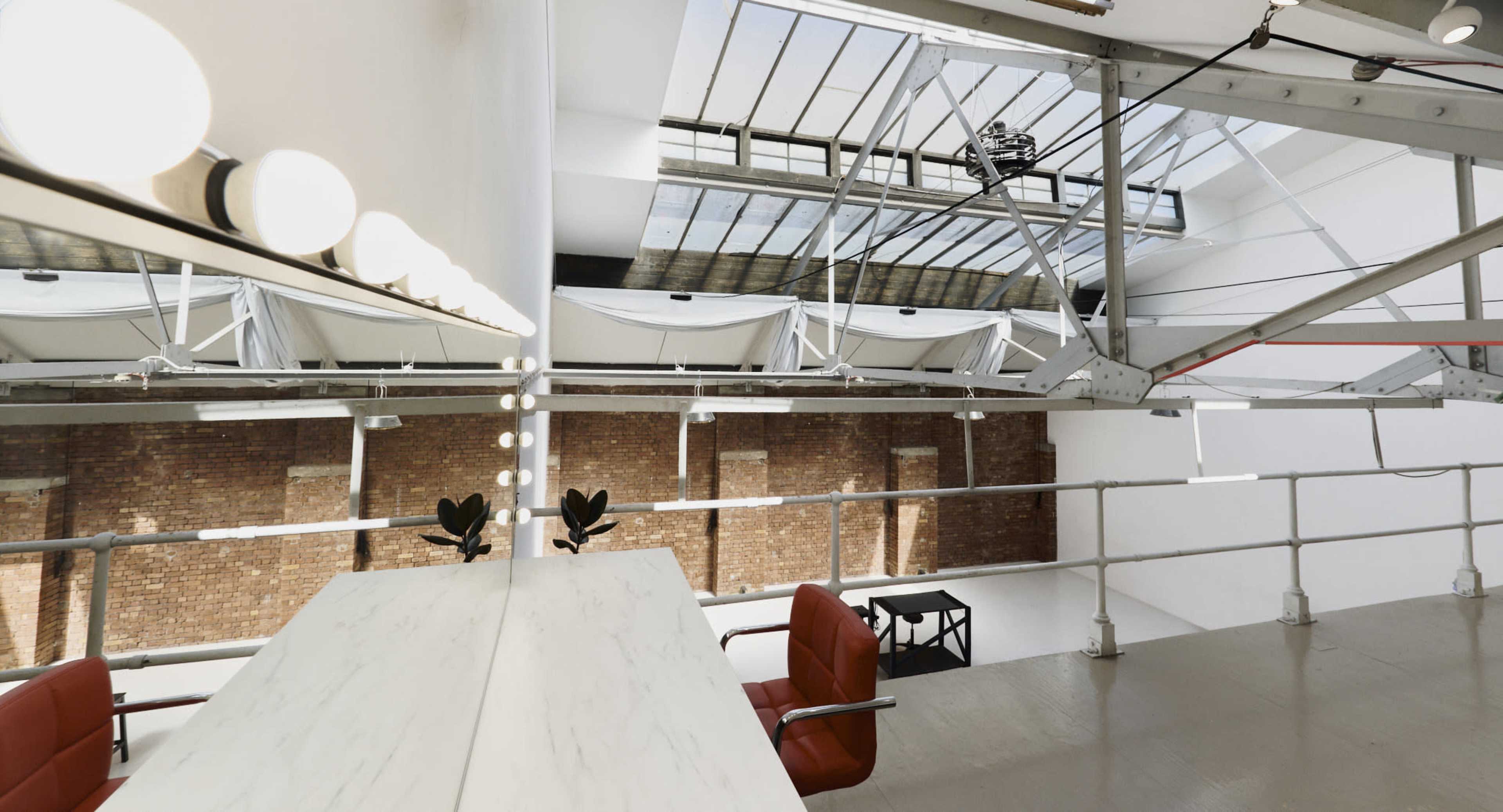 The image shows a spacious, well-lit loft-style interior with exposed brick walls, a modern table, and two red chairs near a vanity with lights.