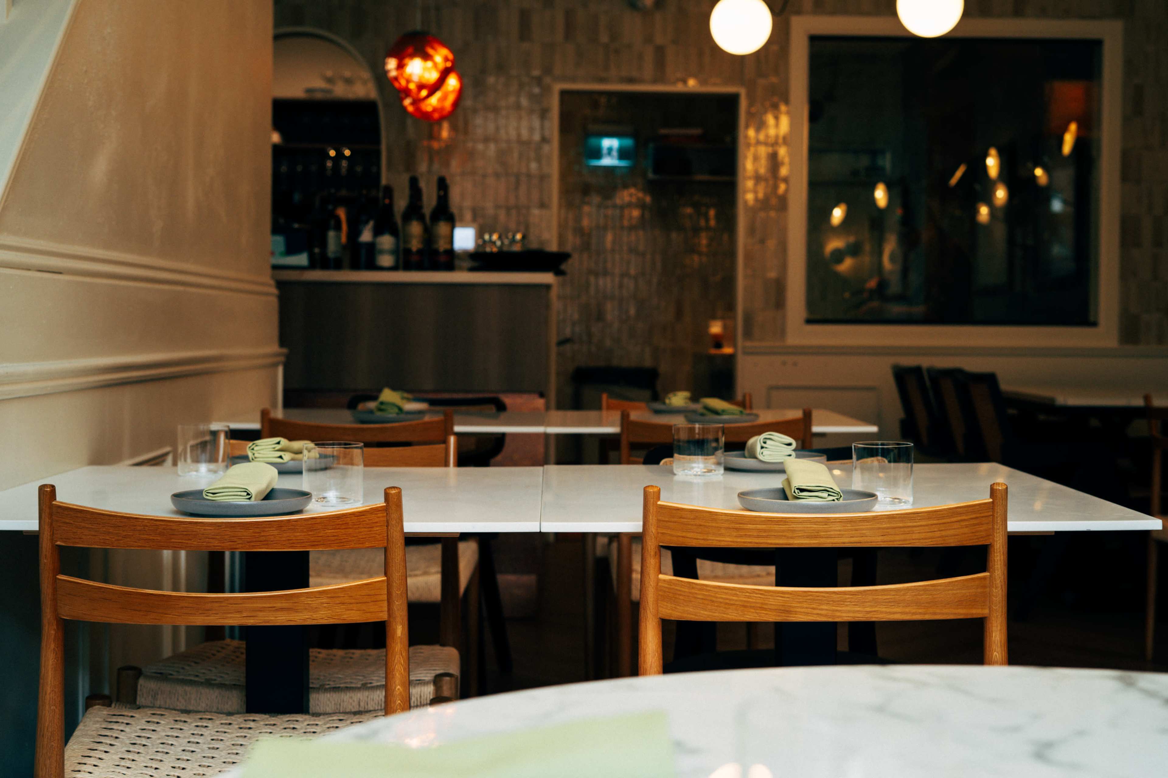 A restaurant interior features empty tables set with plates and glasses, with a bar visible in the background.