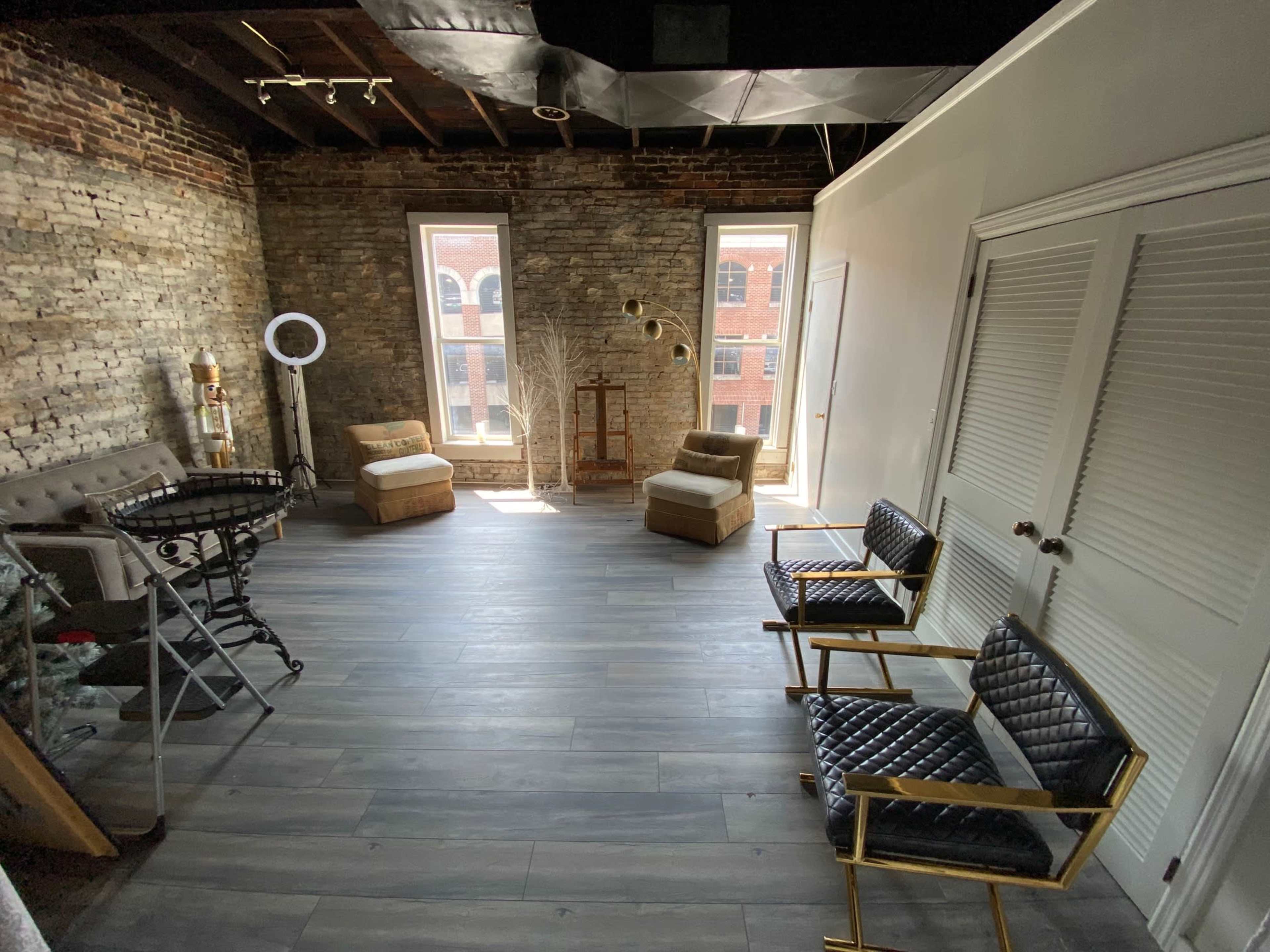 The image shows a room with exposed brick walls, wooden flooring, and two chairs facing a large window, with additional seating and decorative elements arranged nearby.
