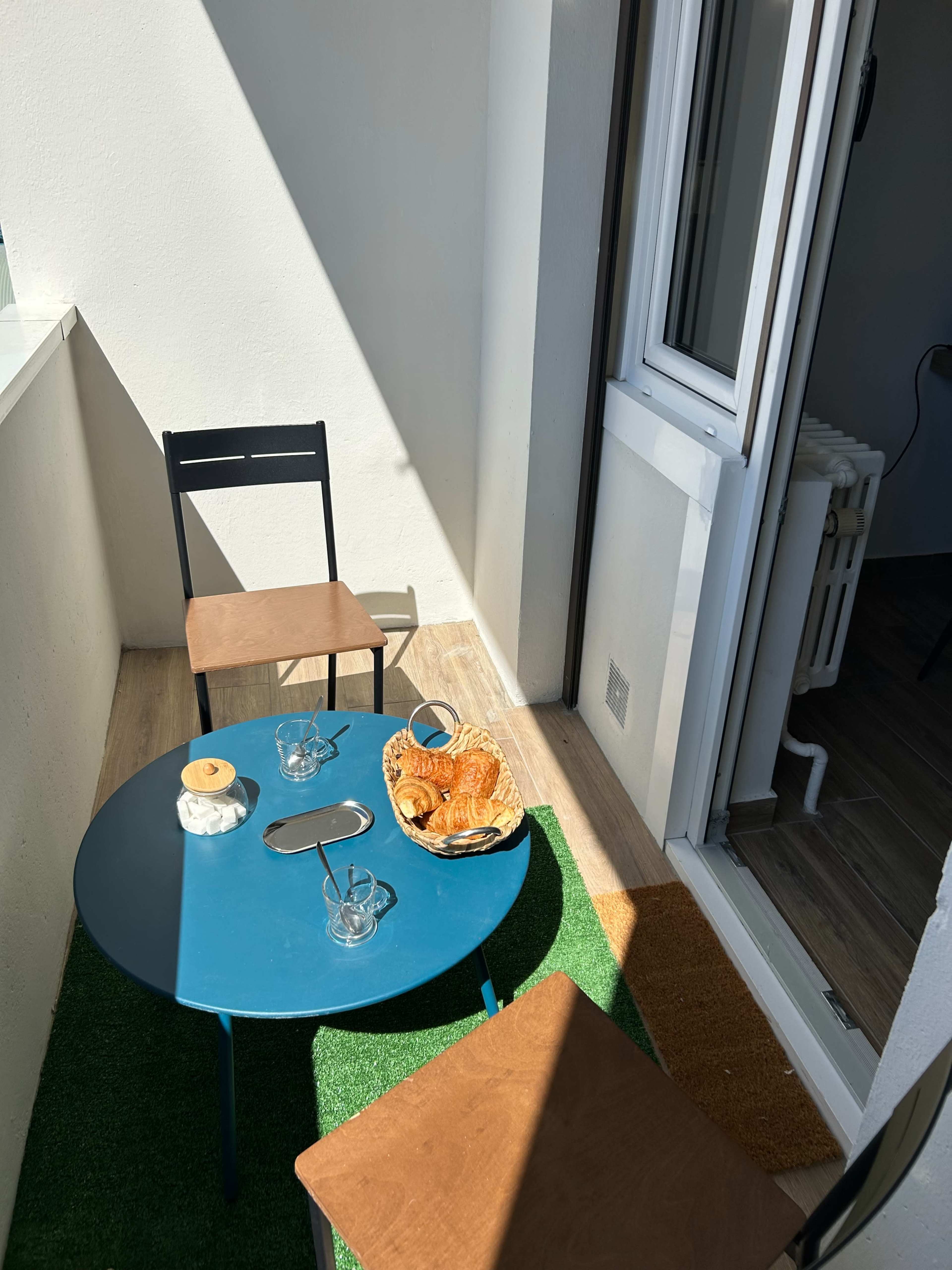 The image shows a small balcony with a round teal table, two chairs, and a plate of pastries on the table, surrounded by artificial grass.
