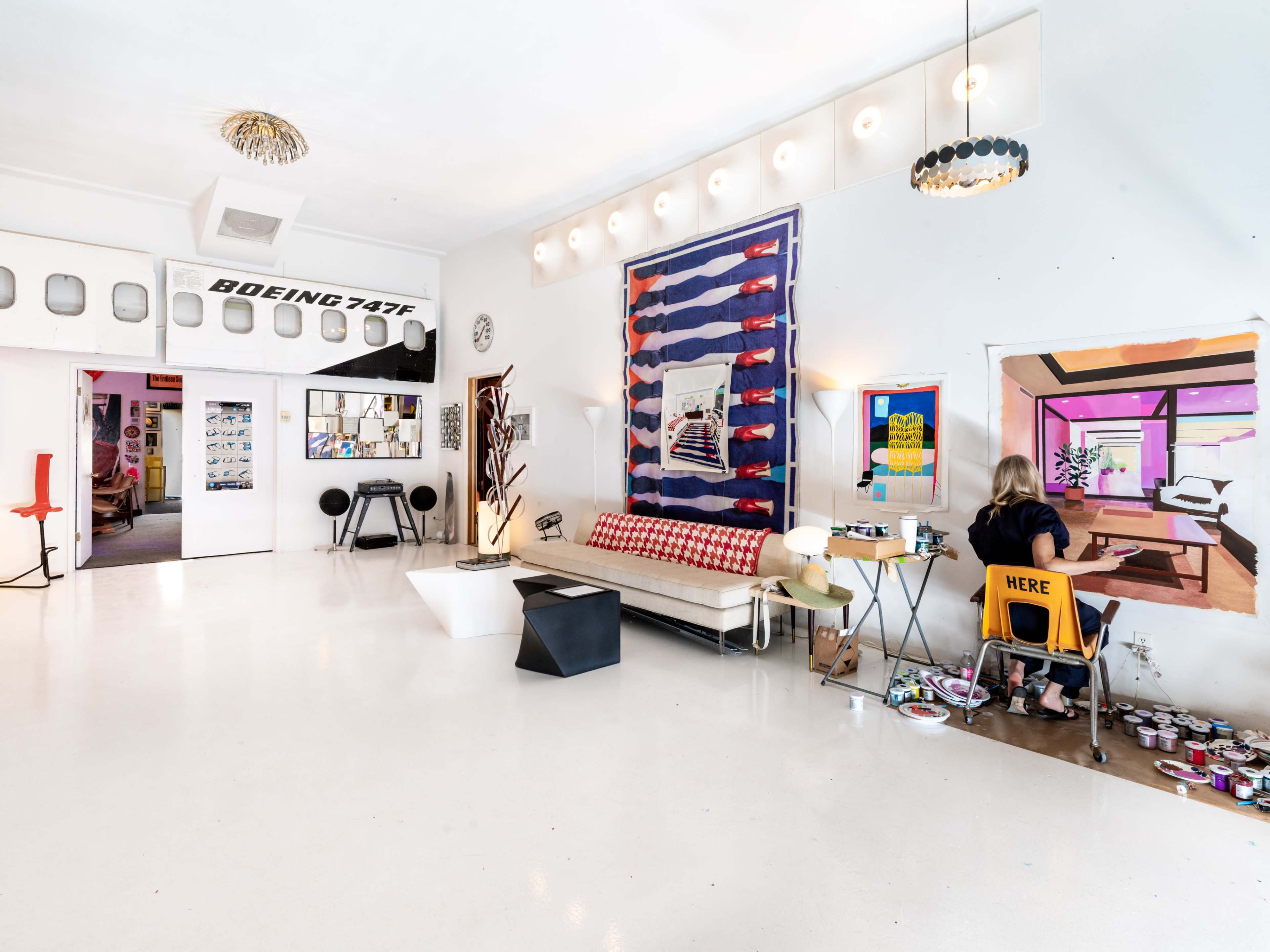 The image shows a bright, open studio space with a modern aesthetic, featuring a central seating area, art on the walls, and a person seated at a small table engaged in creative work.