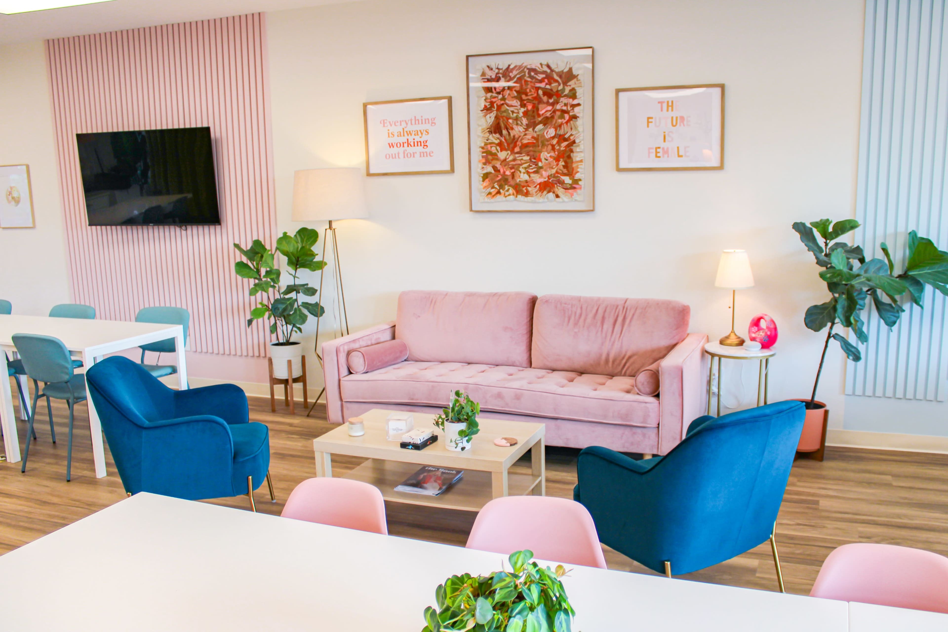 The image shows a modern, stylish living space with a pink sofa, blue chairs, a wooden coffee table, and various plants.