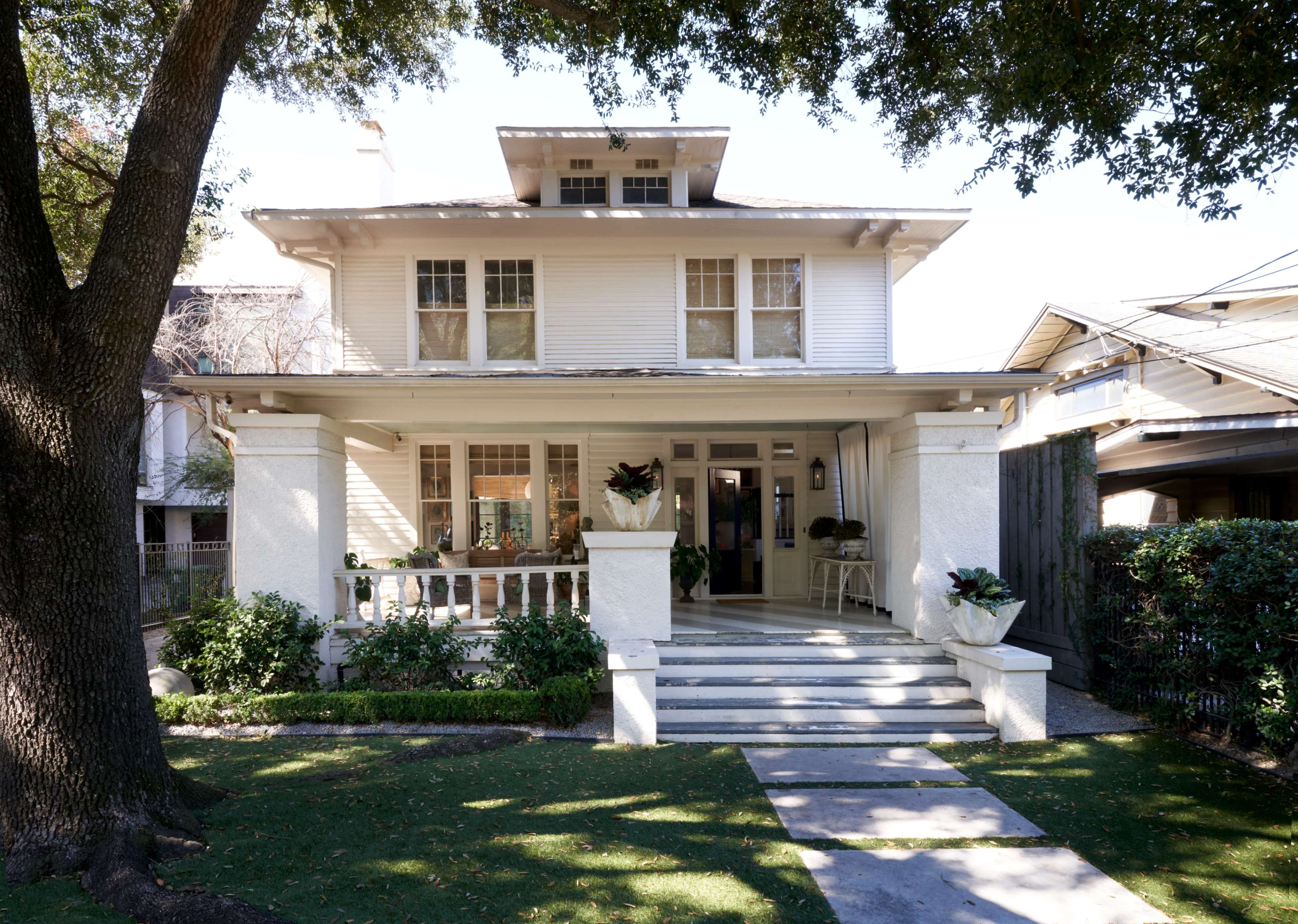 A two-story white house with a porch, surrounded by greenery and a pathway leading to the entrance.