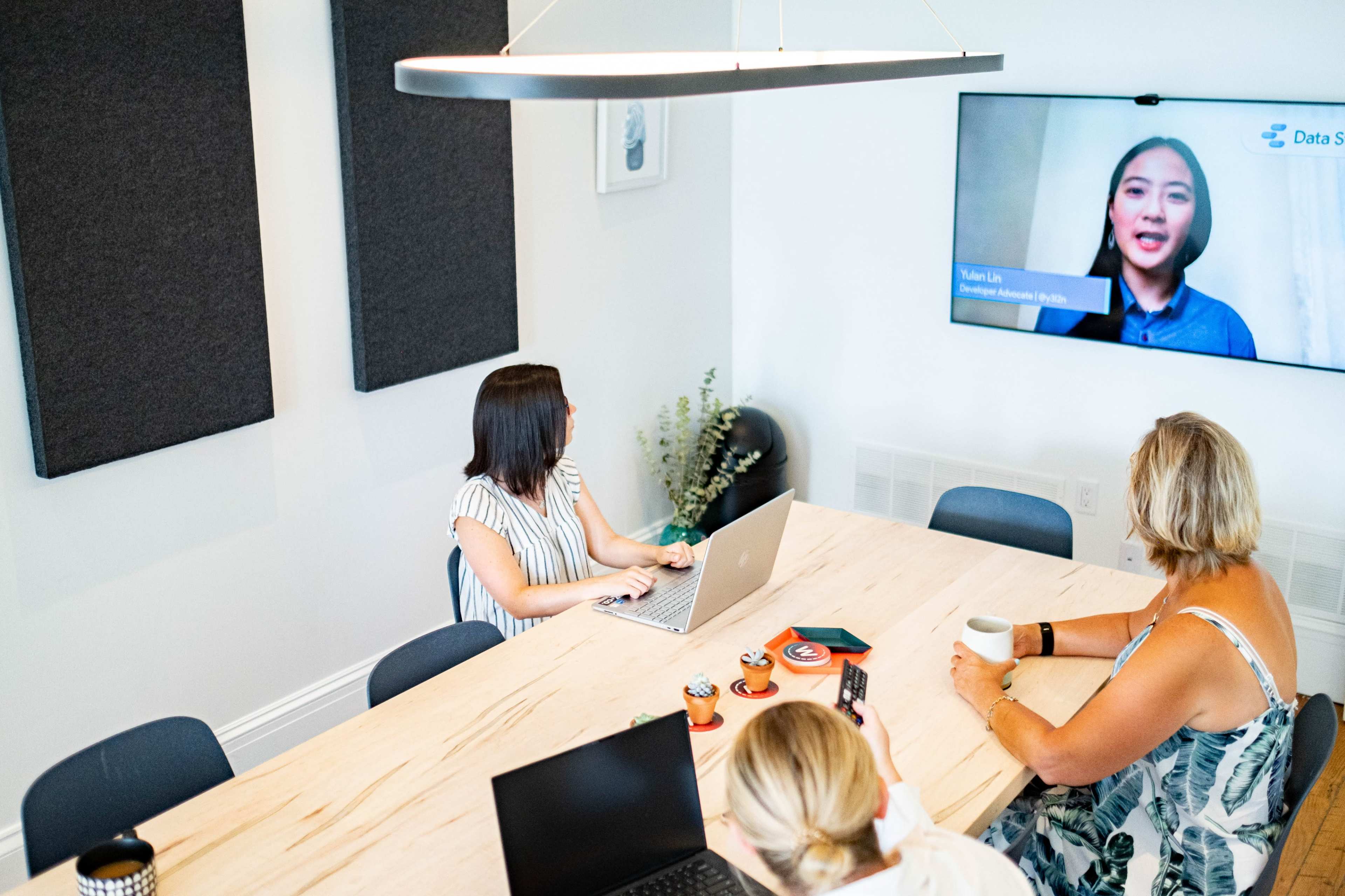 A group of three people are sitting around a wooden table in a conference room, with one participant visible on a screen while another works on a laptop.