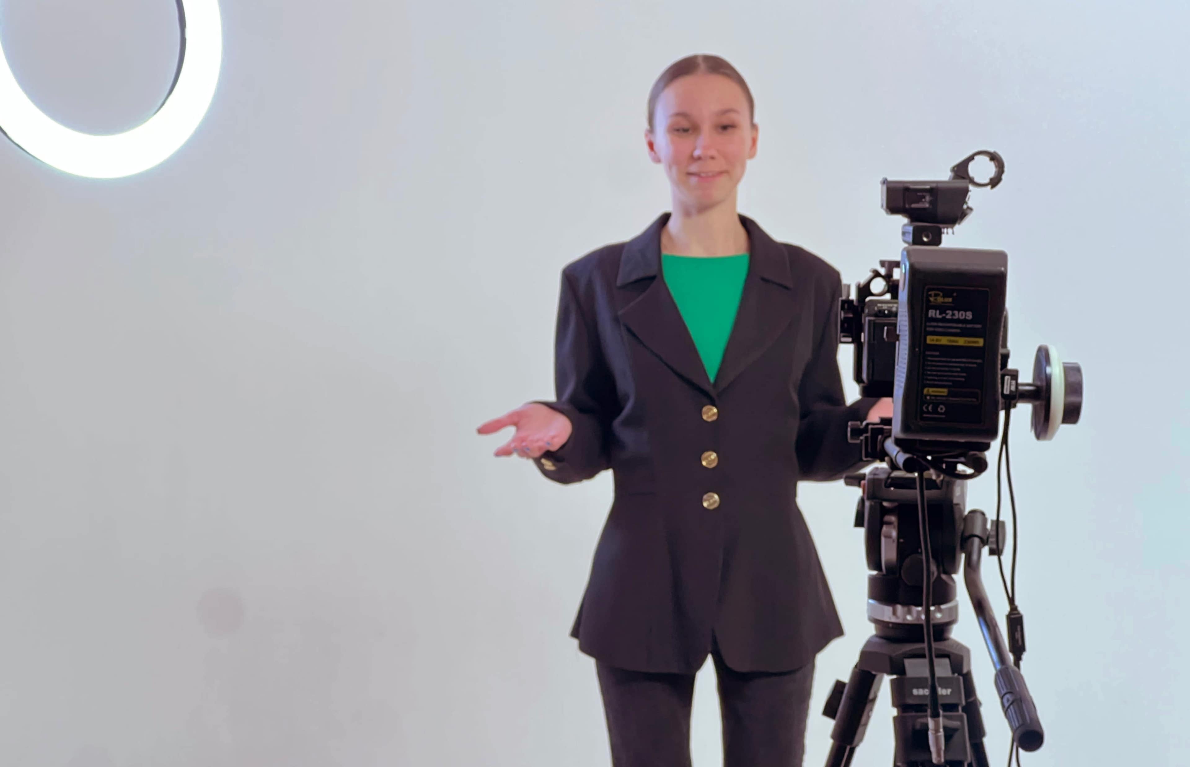 A person in a blazer stands next to a camera on a tripod against a light background.