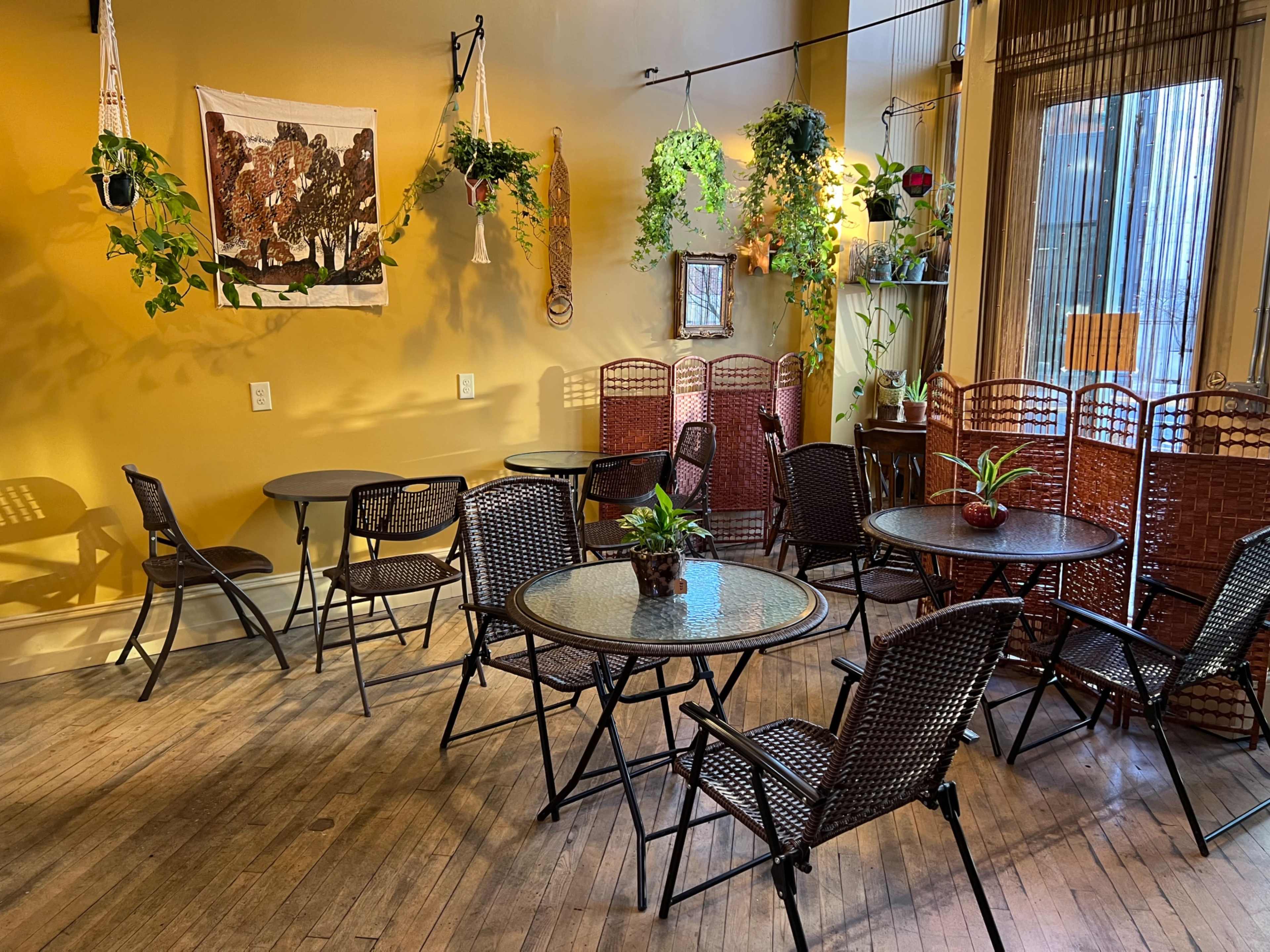 A cozy café space with several round tables and chairs, adorned with plants and a decorative wall hanging.