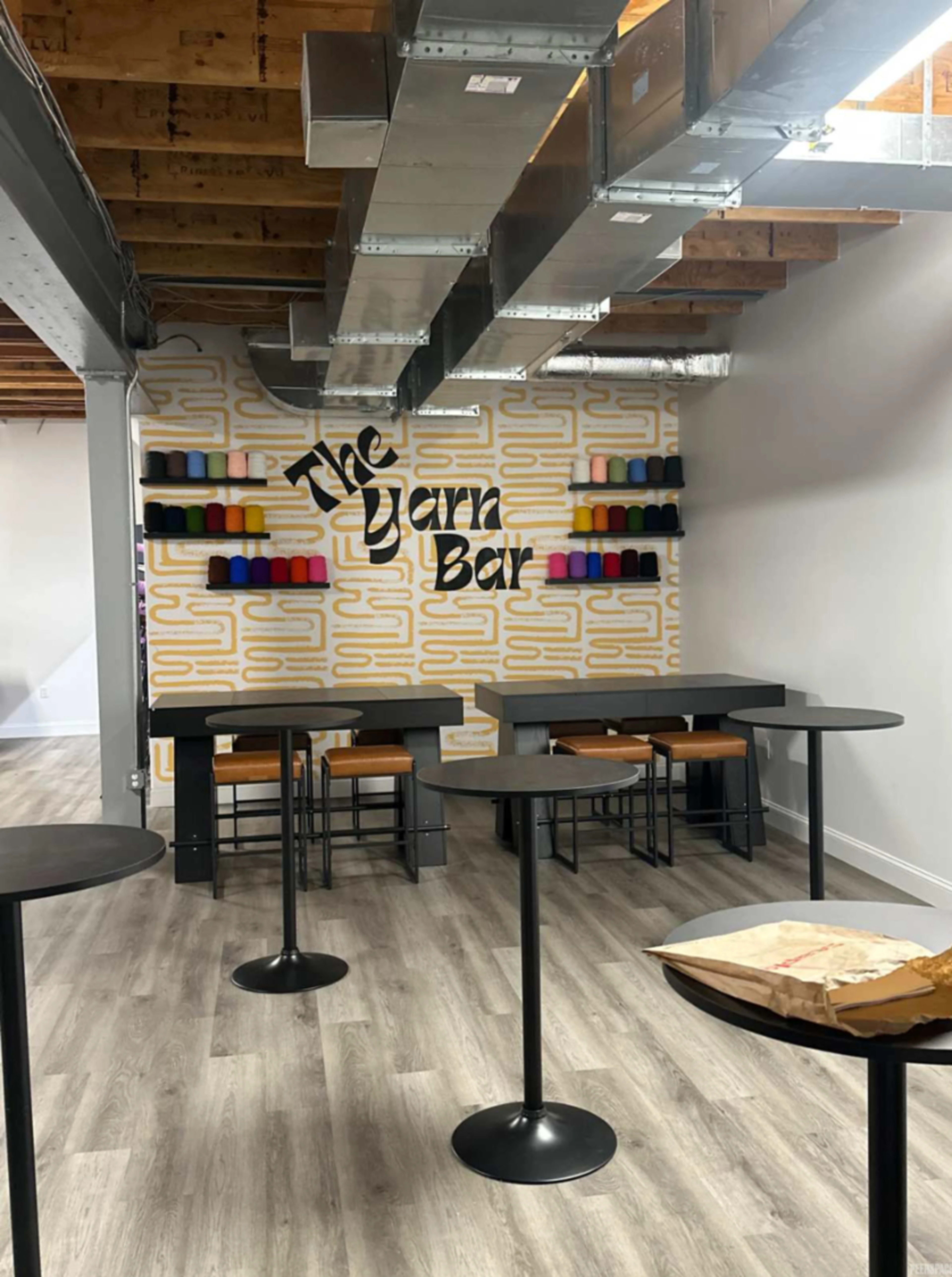 The image shows a modern interior space called "The Yarn Bar," featuring a patterned wall, several shelves with colorful yarn spools, and a few black tables and stools arranged around the area.