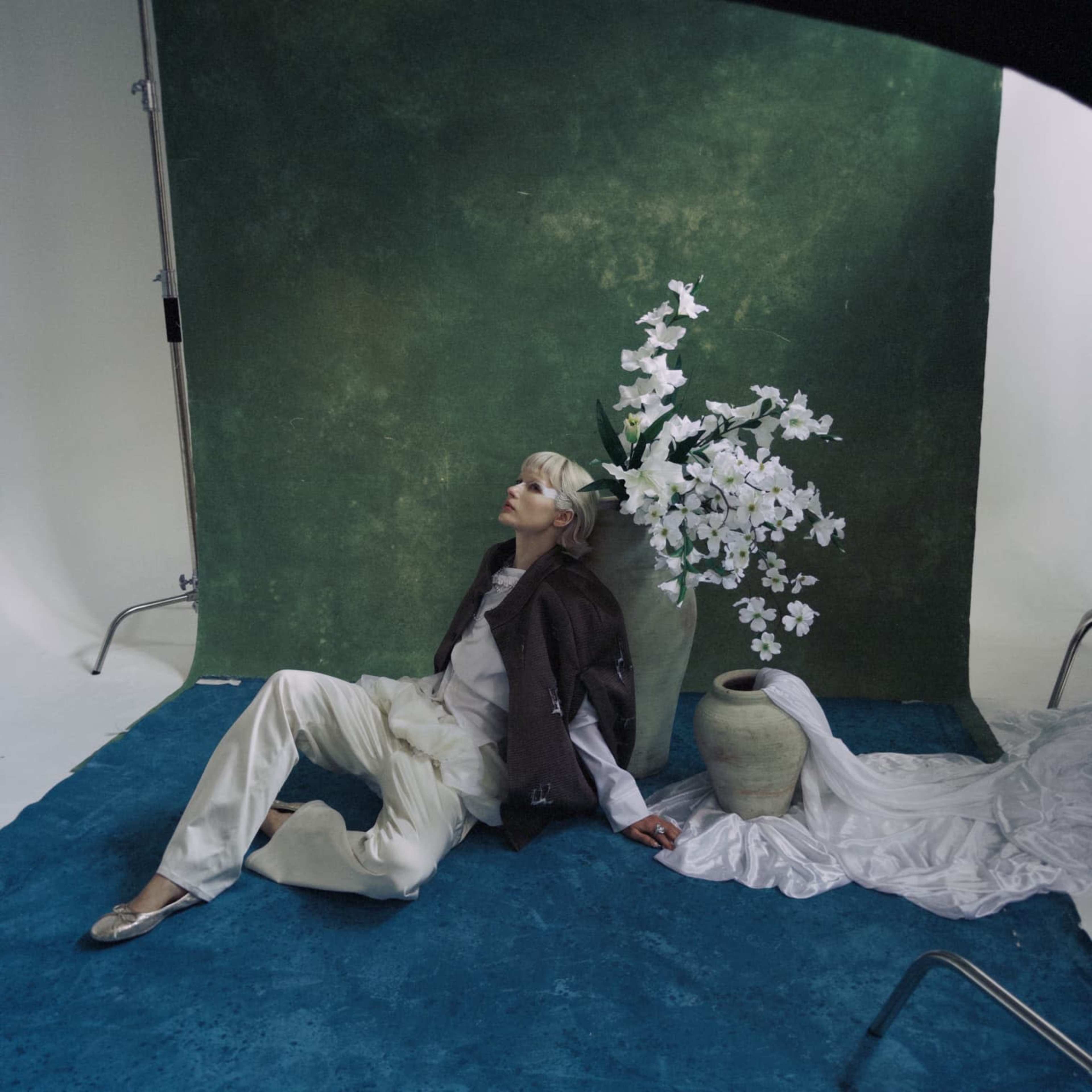 A model in a stylish outfit poses beside a large vase filled with white flowers against a green backdrop in a studio setting.