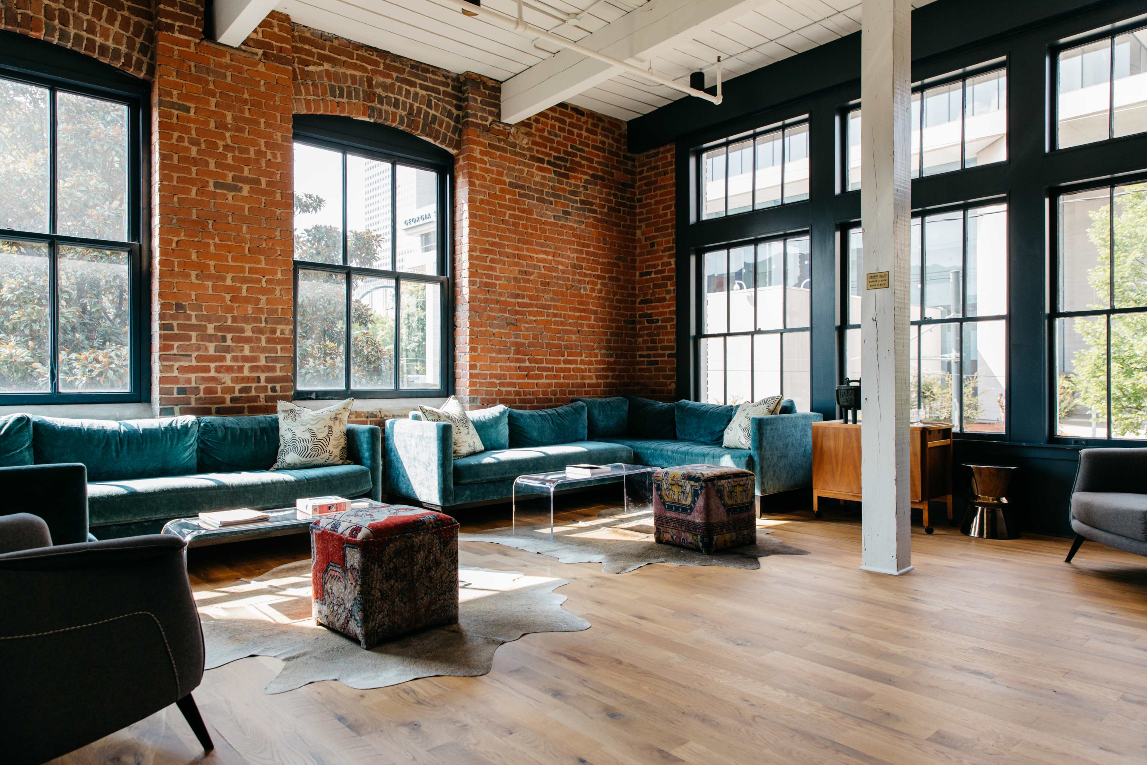 A spacious lounge area features a large sectional sofa, a cowhide rug, and various seating options against a backdrop of exposed brick walls and large windows.