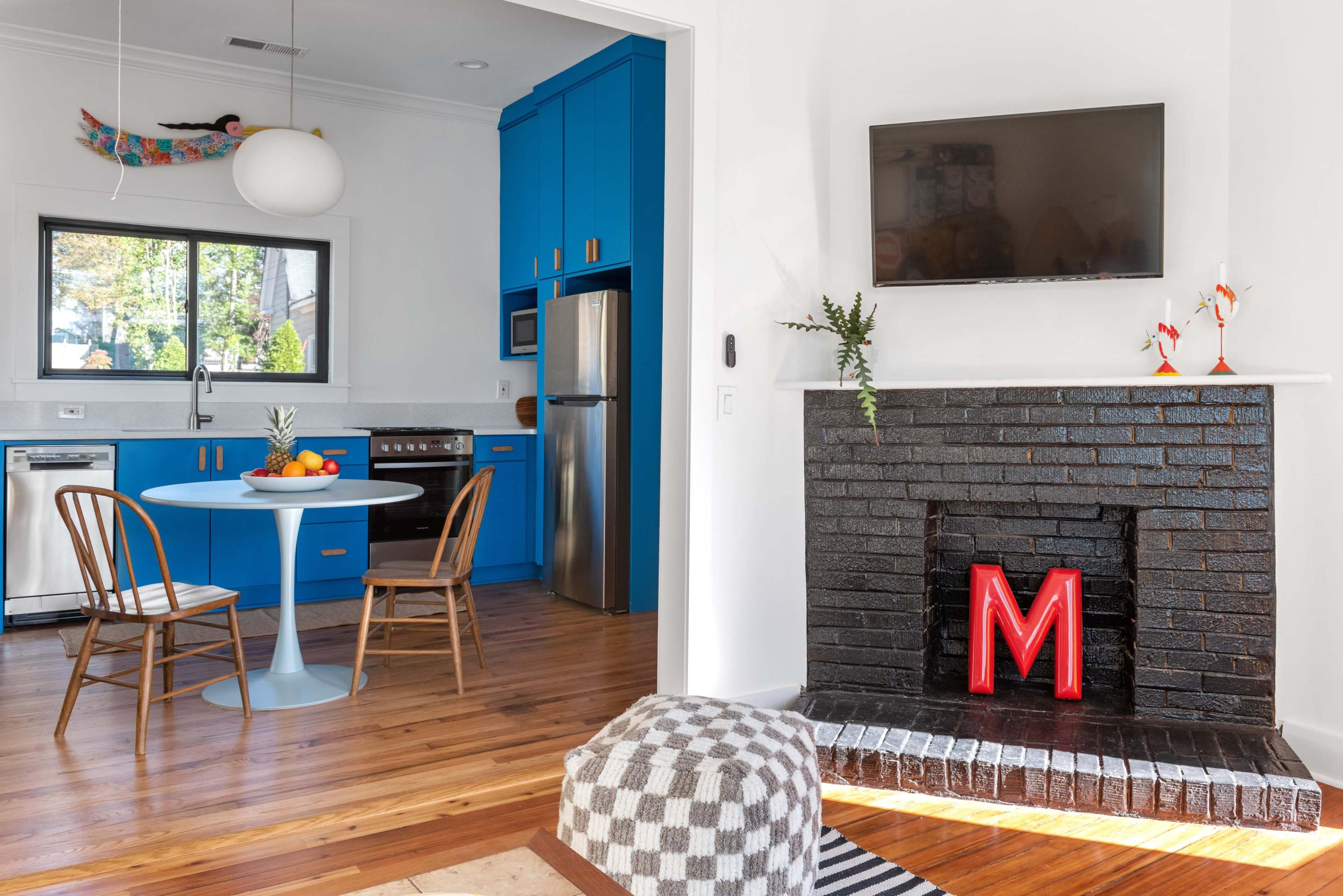 The image shows a modern kitchen with blue cabinetry, a round table with two chairs, and a living area featuring a black brick fireplace with a red letter "M" displayed in front.