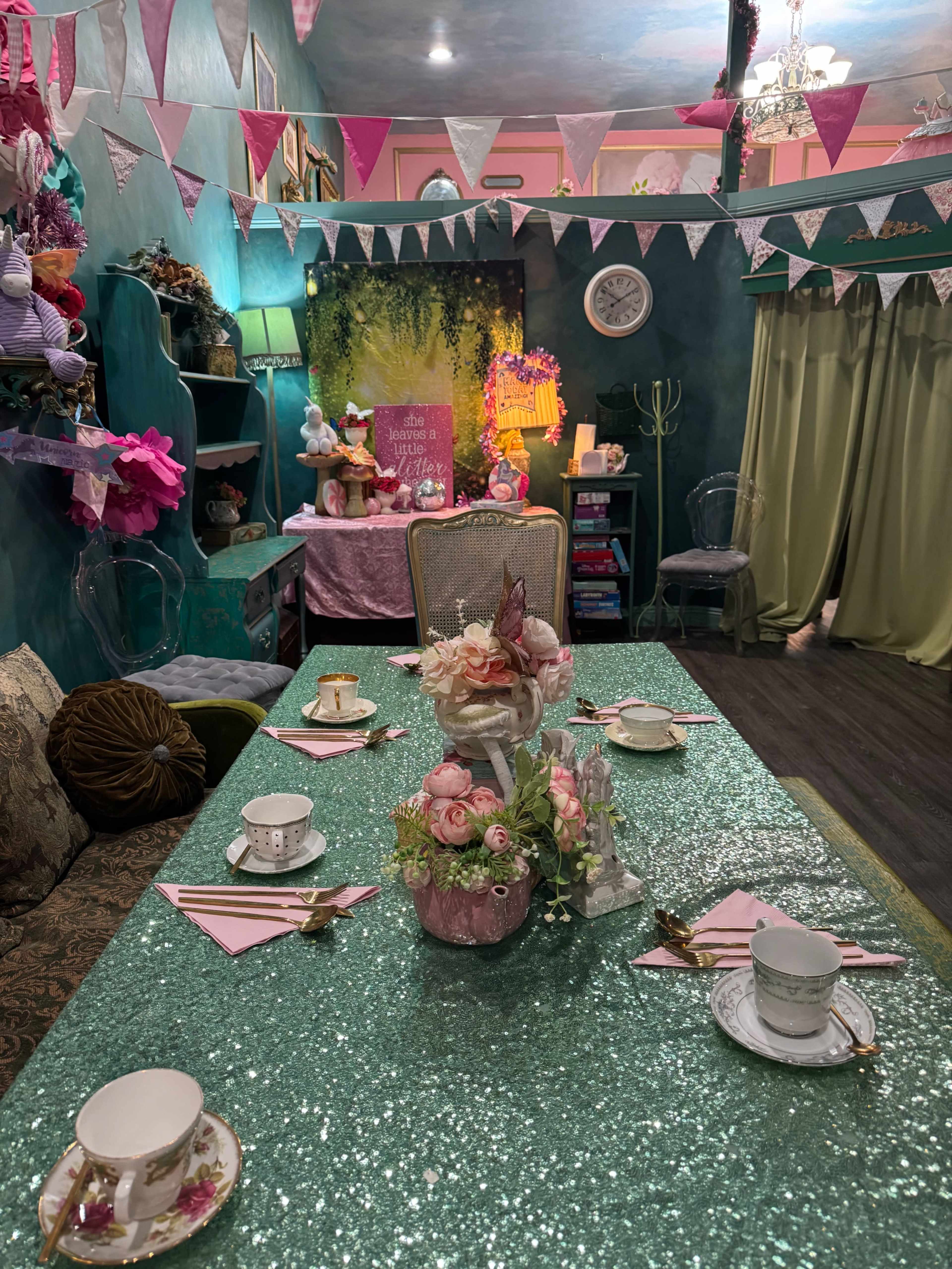The image shows a brightly decorated tea party setup with a sparkling green tablecloth, floral centerpieces, and elegant tea cups, surrounded by colorful decor and bunting.