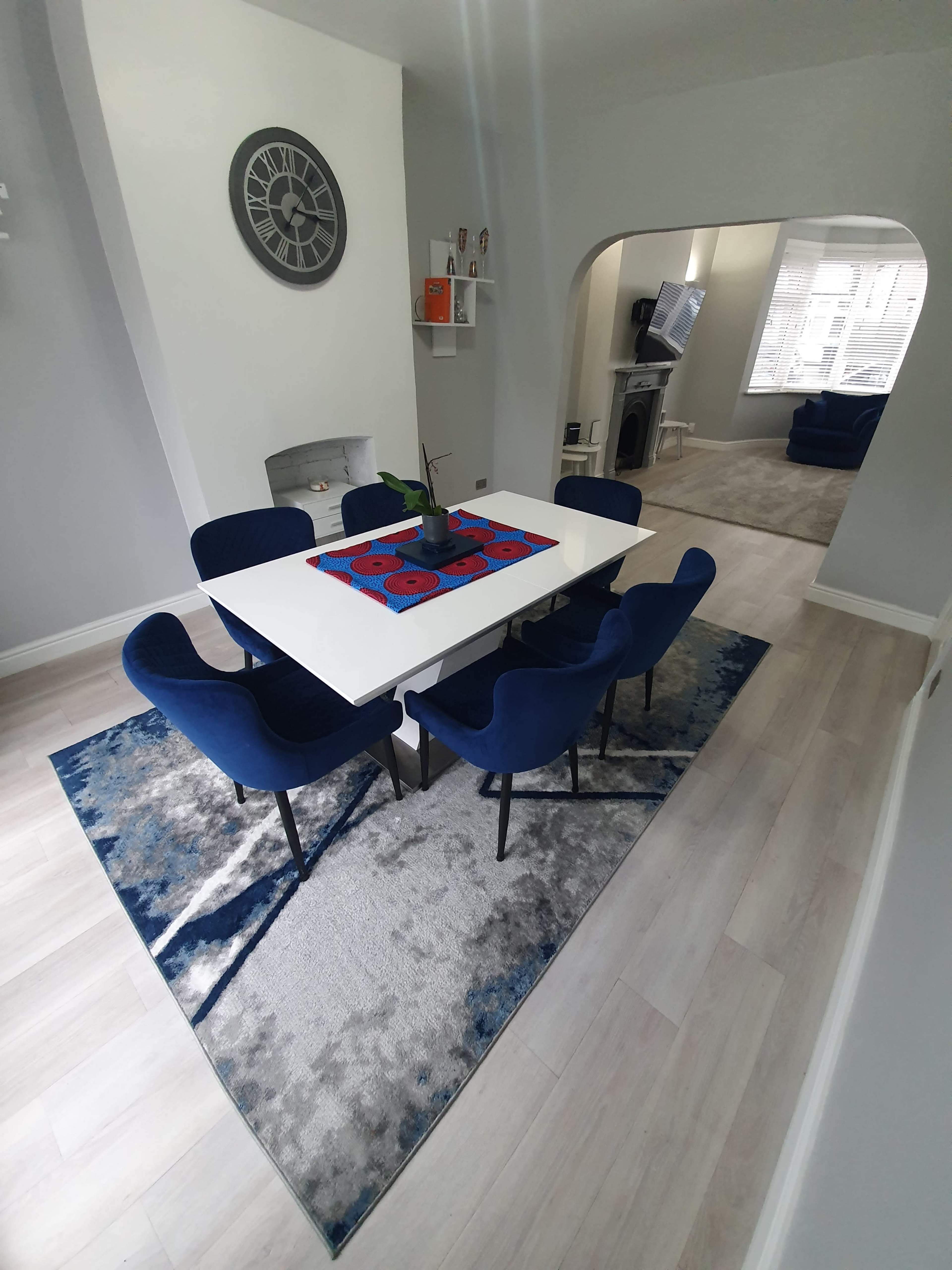 A dining area with a white table surrounded by blue chairs, a patterned rug on the floor, and an archway leading to a living room space.