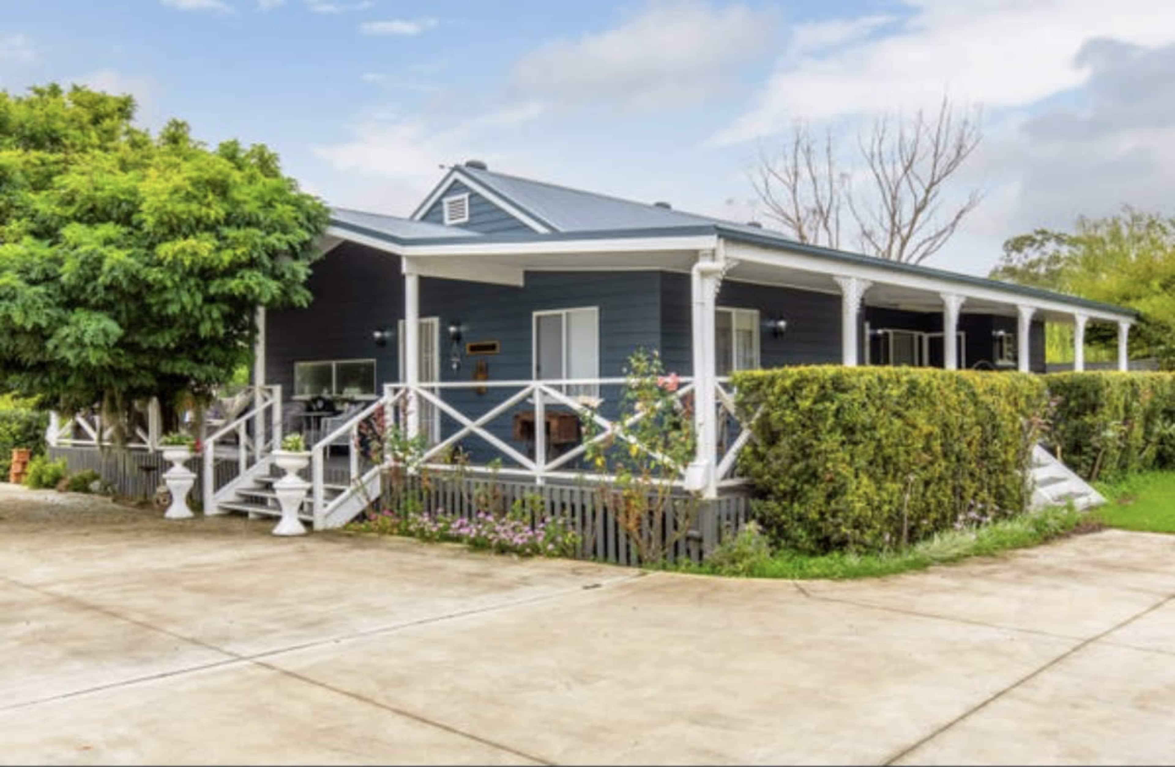 A blue house with a front porch and surrounding greenery is situated on a concrete driveway.