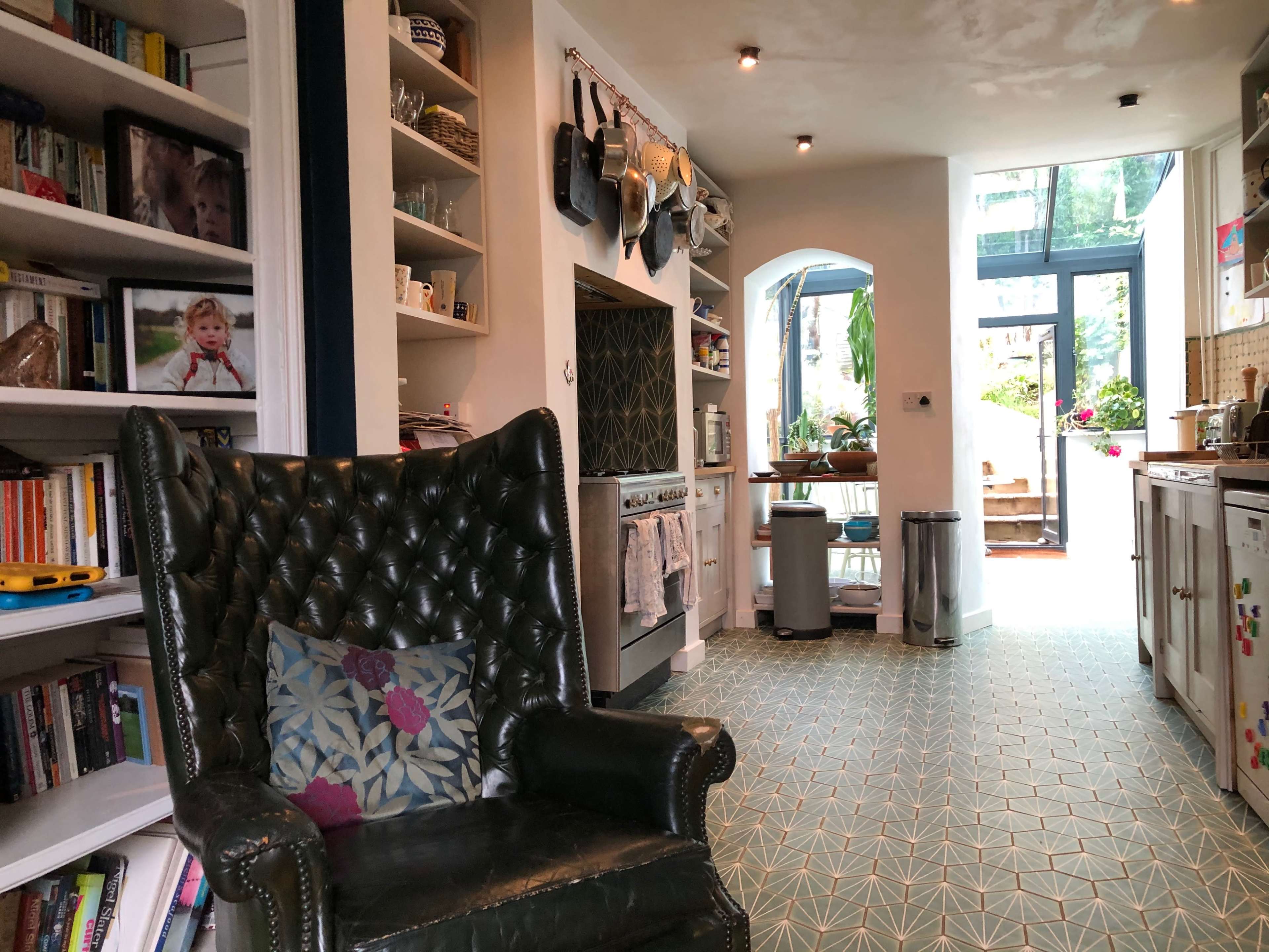The image shows a cozy kitchen area featuring a green upholstered armchair, shelves filled with books and photos, and a view of a backyard through large windows.
