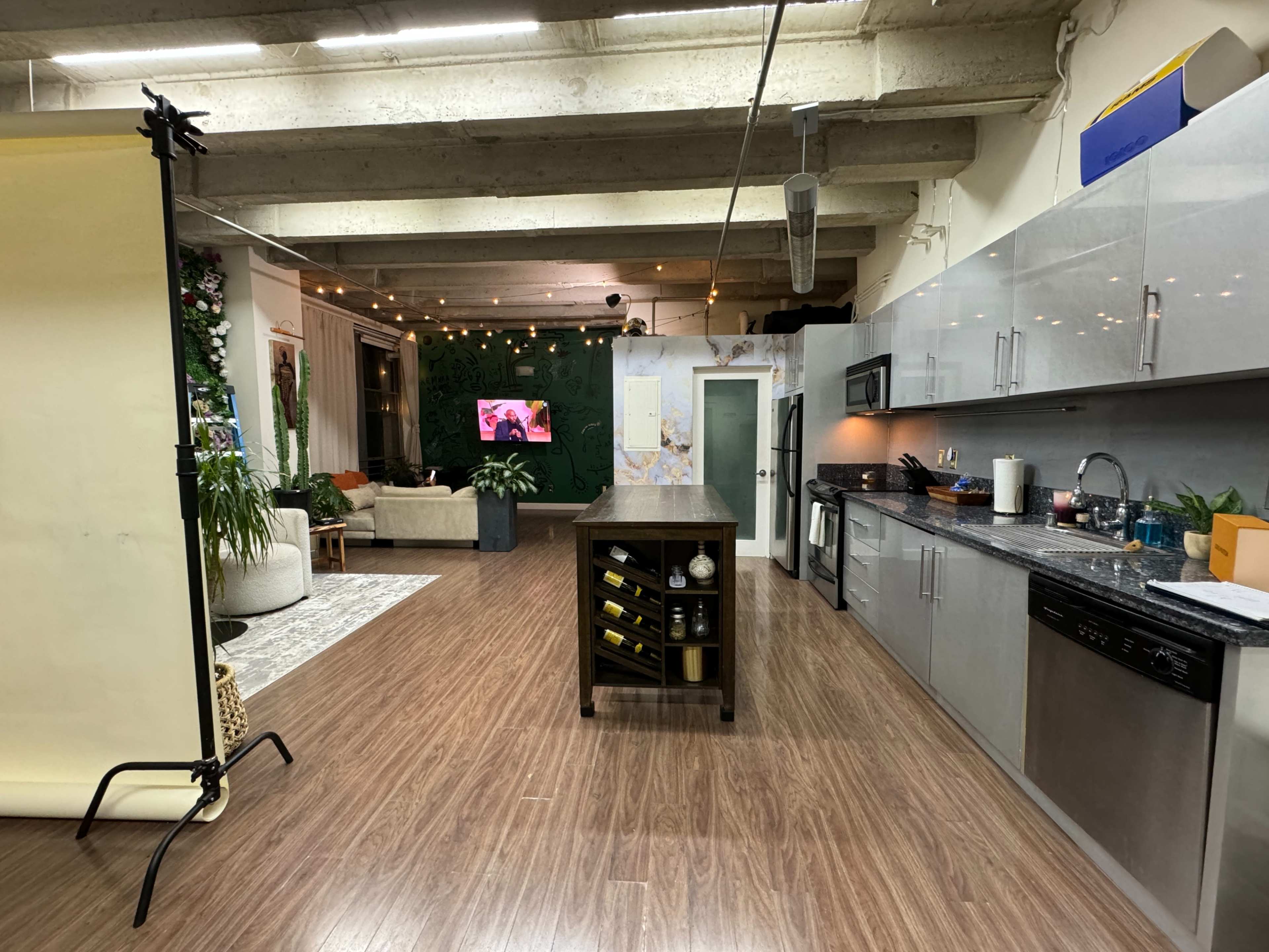 A spacious kitchen area with modern appliances, an island with wine storage, and a living room visible in the background decorated with plants and a television on the wall.