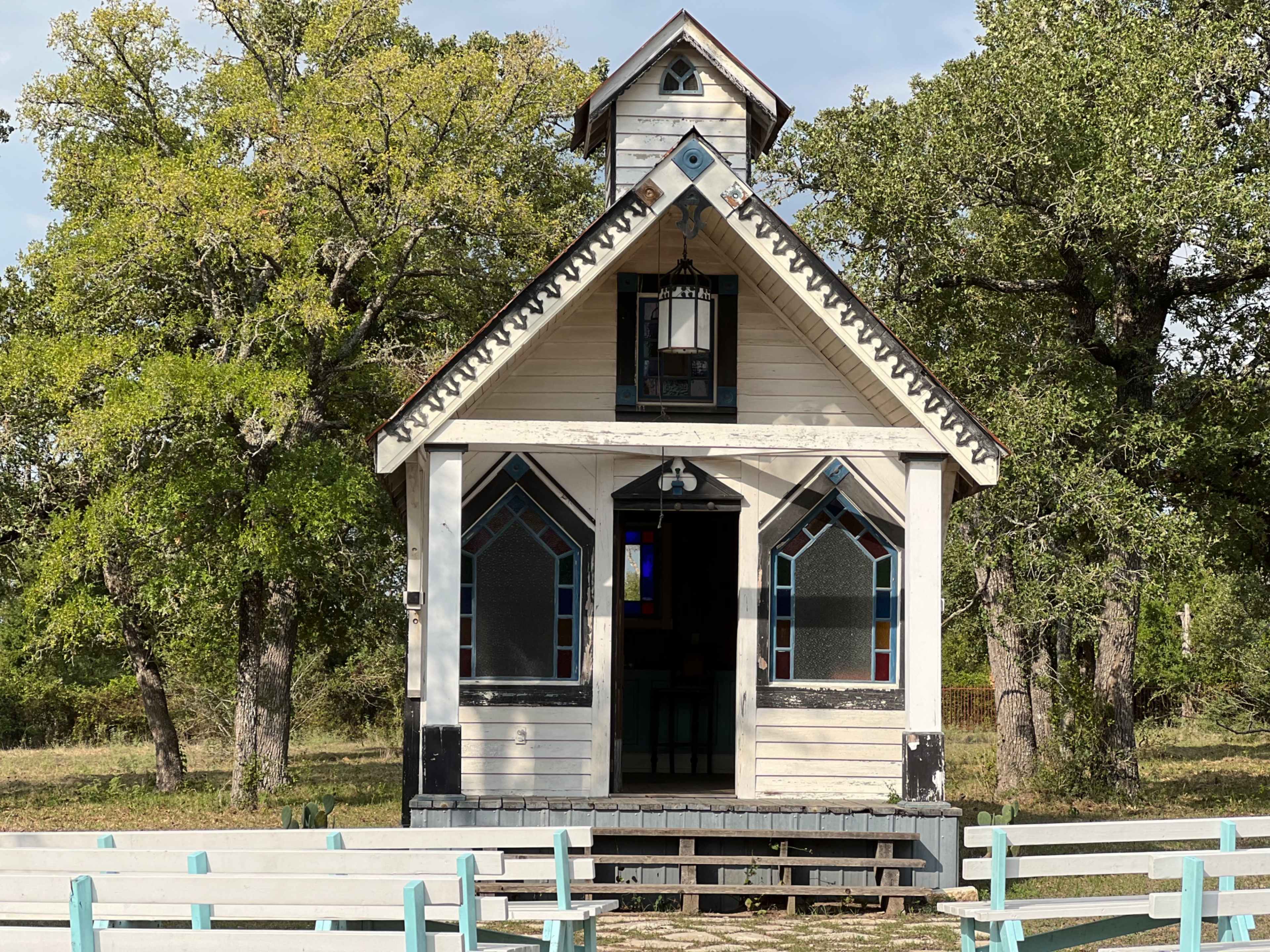 A small, vintage chapel with stained glass windows is situated among trees, surrounded by a white railing fence.
