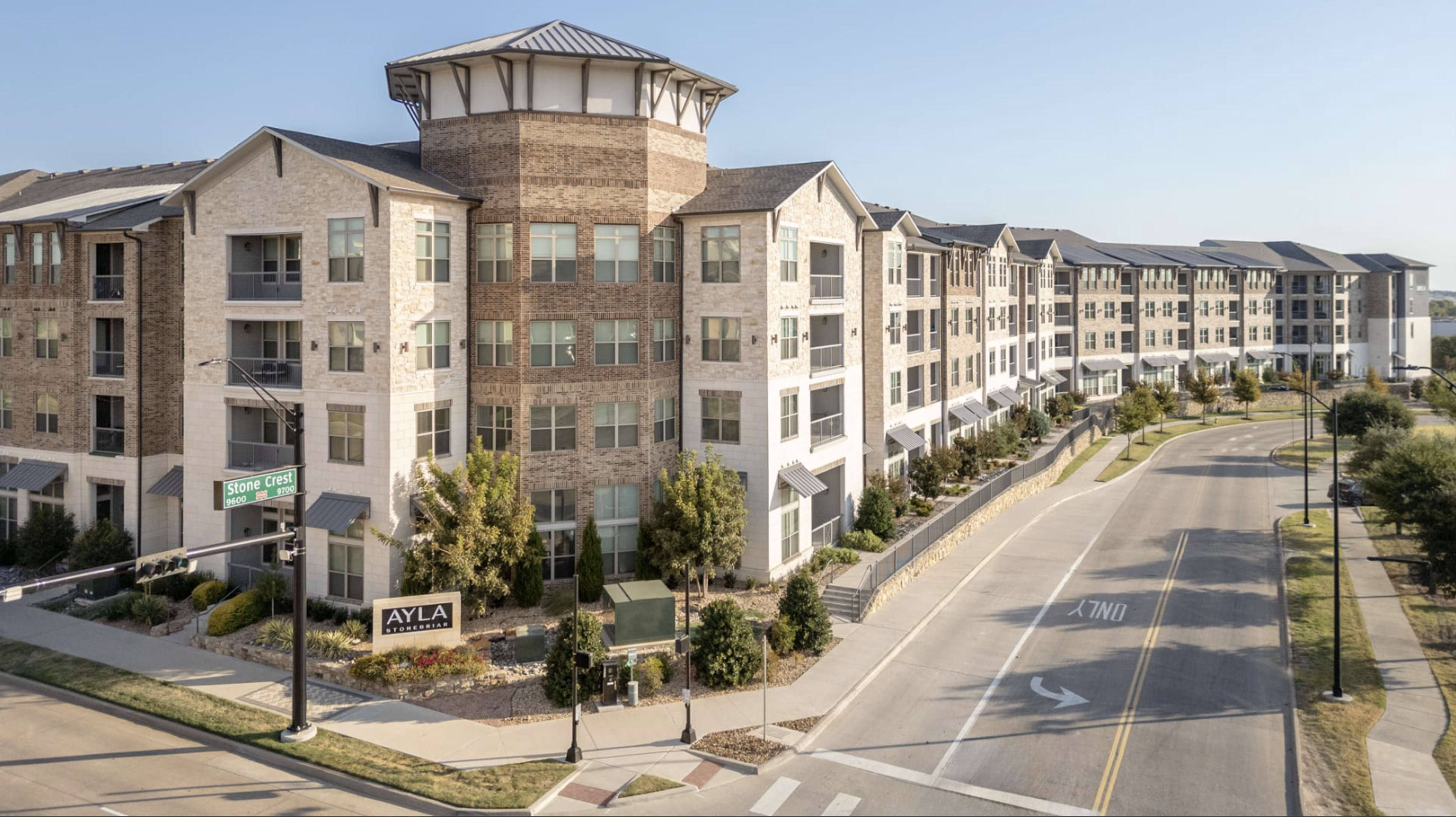 The image shows a multi-story apartment complex with a unique architectural design, situated along a clean, well-maintained road with landscaped greenery.