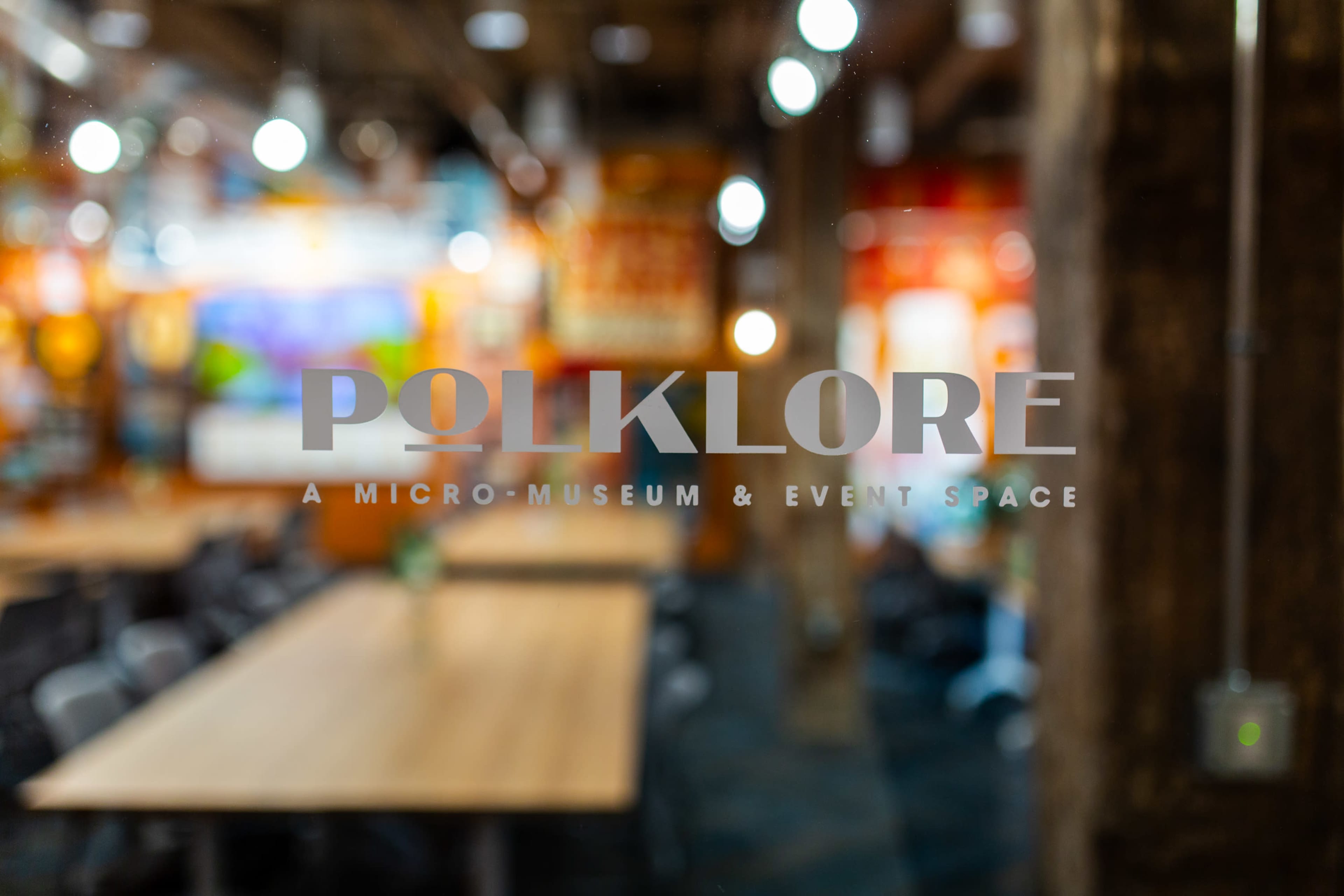 The image shows the sign for "Polklore," identifying it as a micro-museum and event space, with a blurred interior of the venue visible in the background.