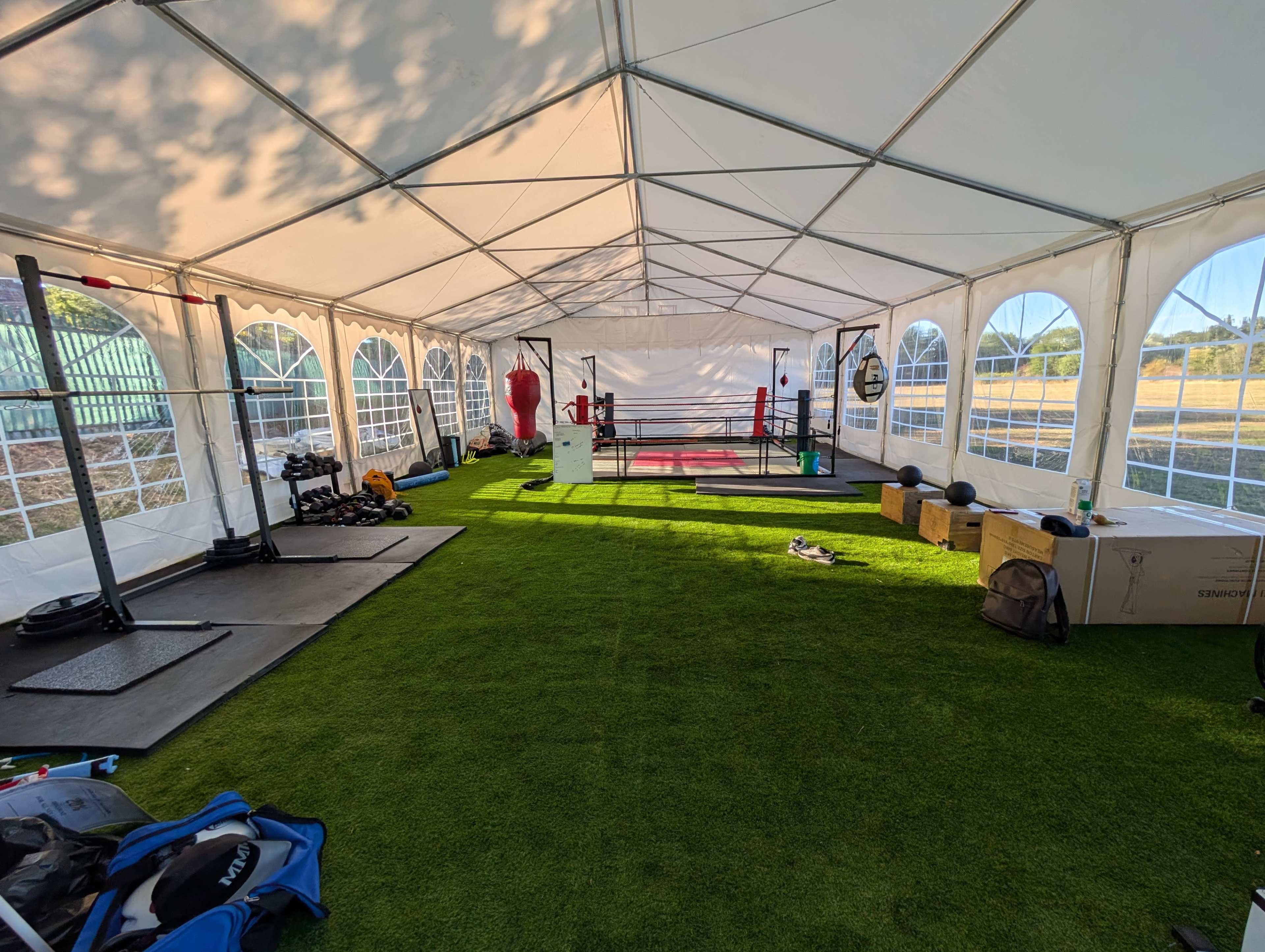 The image shows the interior of a large tent set up as a training facility, featuring various workout equipment, a boxing ring, and artificial grass flooring.