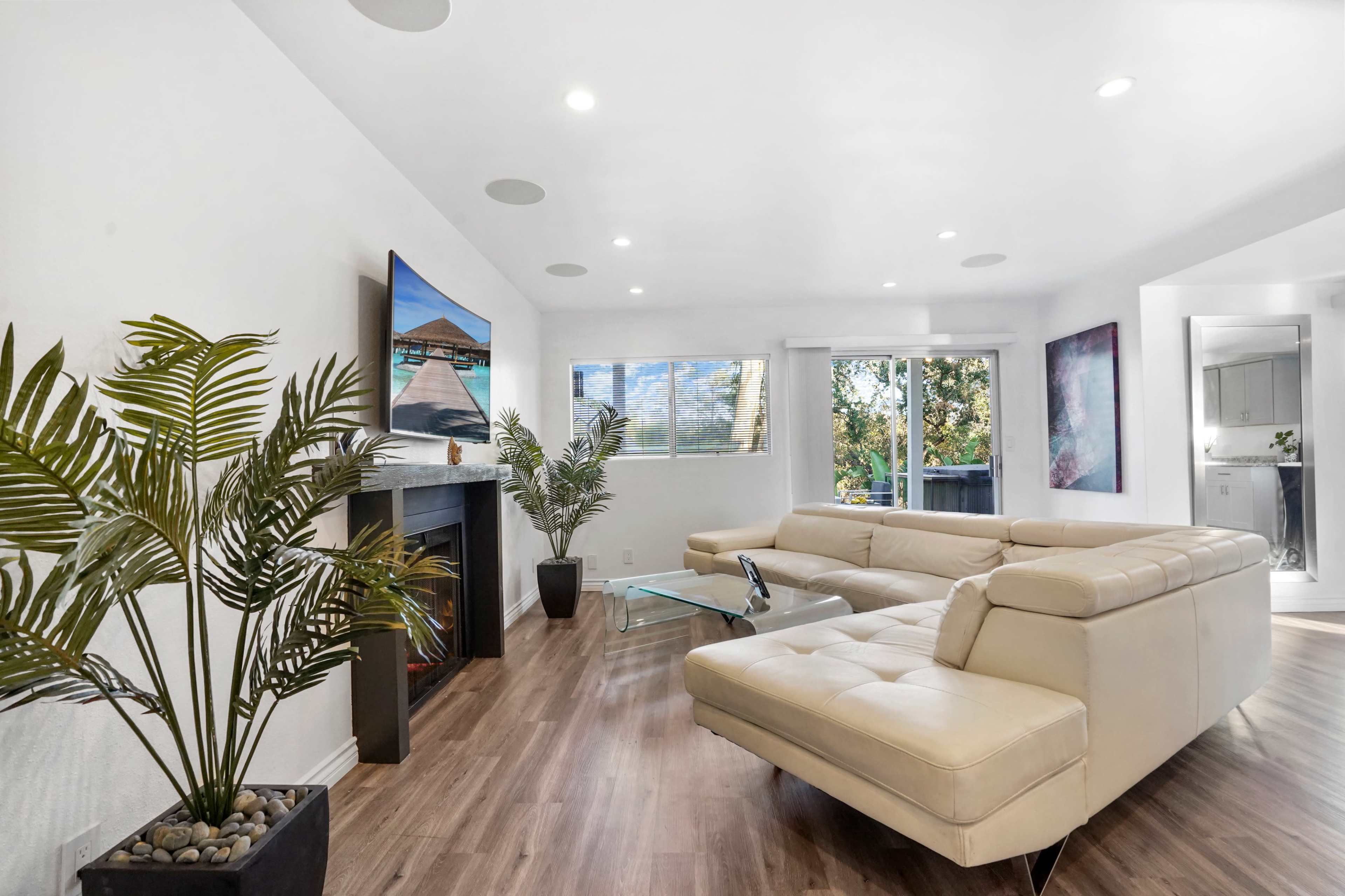 A modern living room with a light-colored sectional sofa, a flat-screen TV mounted on the wall, large windows, and a decorative plant in a pot.
