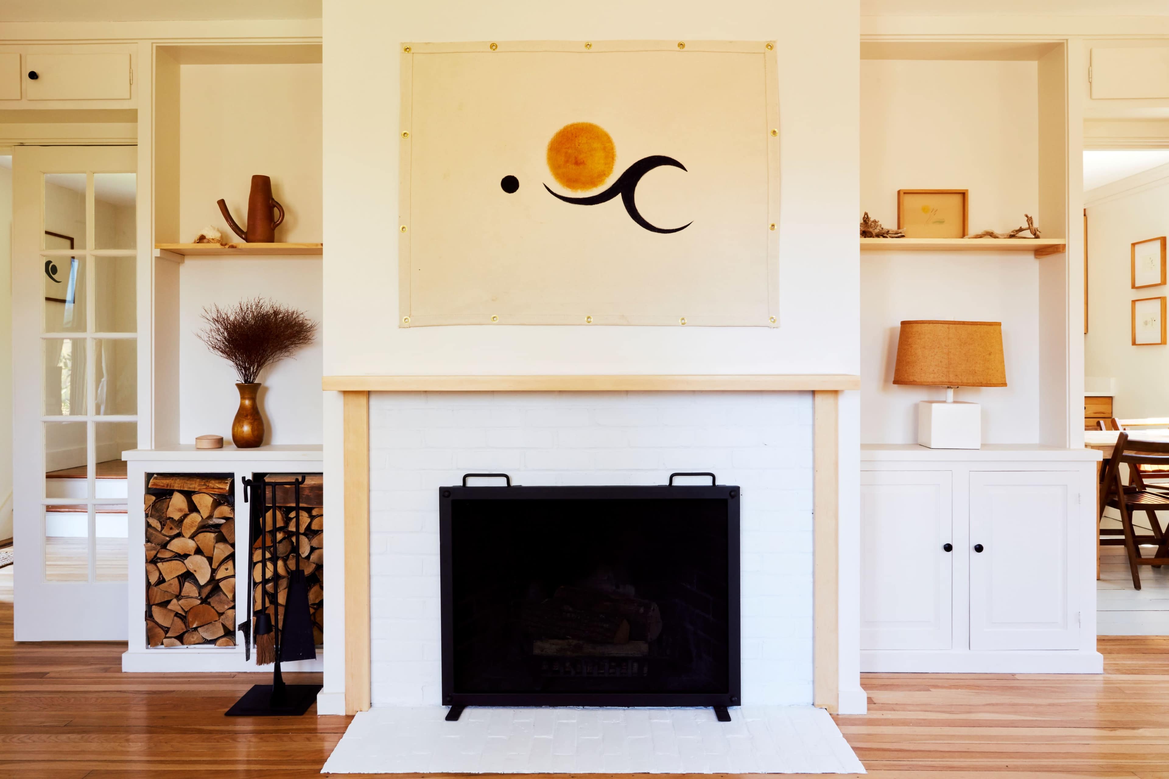 A living room with a white brick fireplace, a wood log storage unit, and a large decorative wall hanging featuring a sun and abstract design.