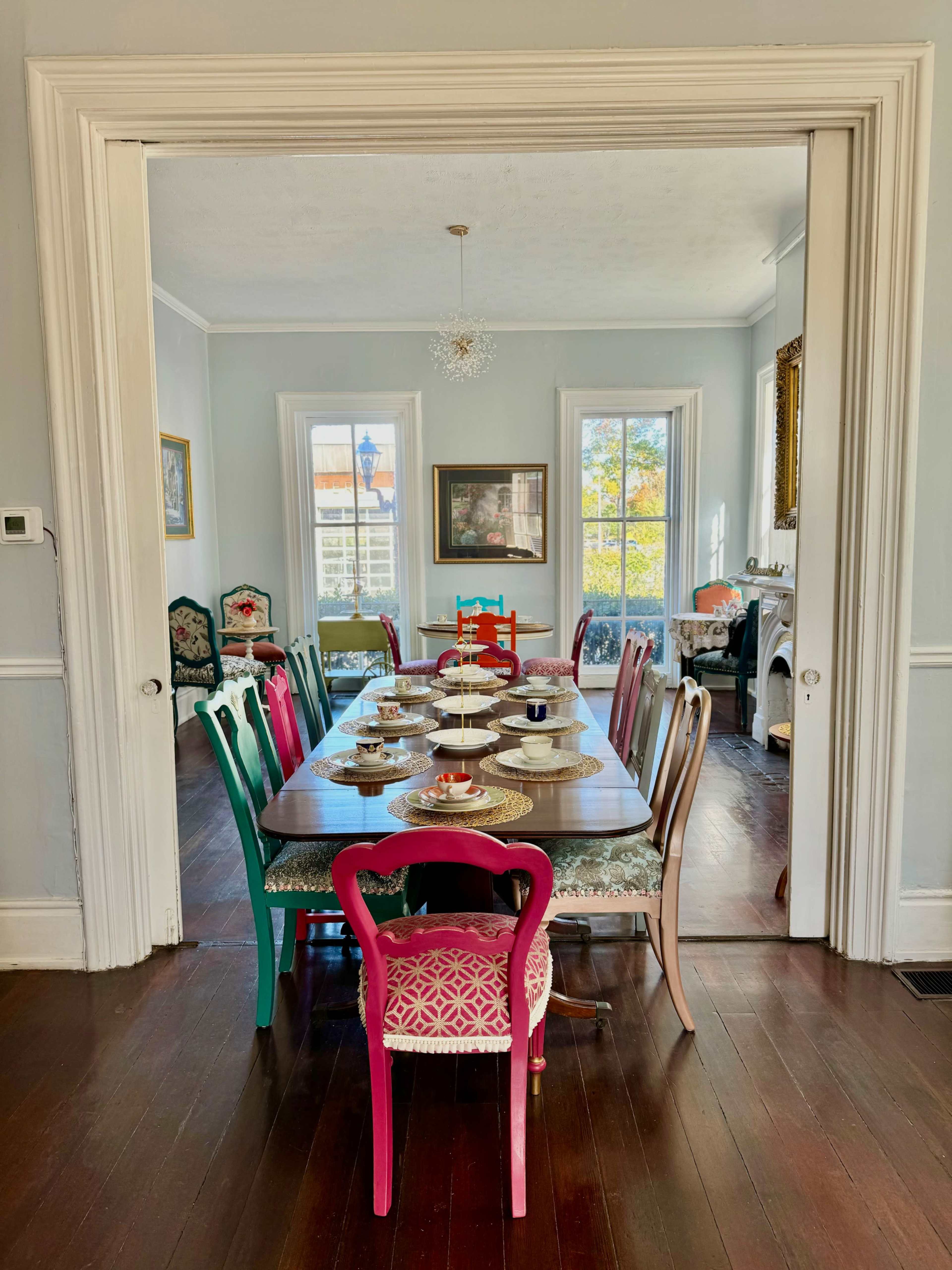 A long dining table is set with plates and cups, surrounded by colorful chairs in a bright, airy room with windows and artwork.