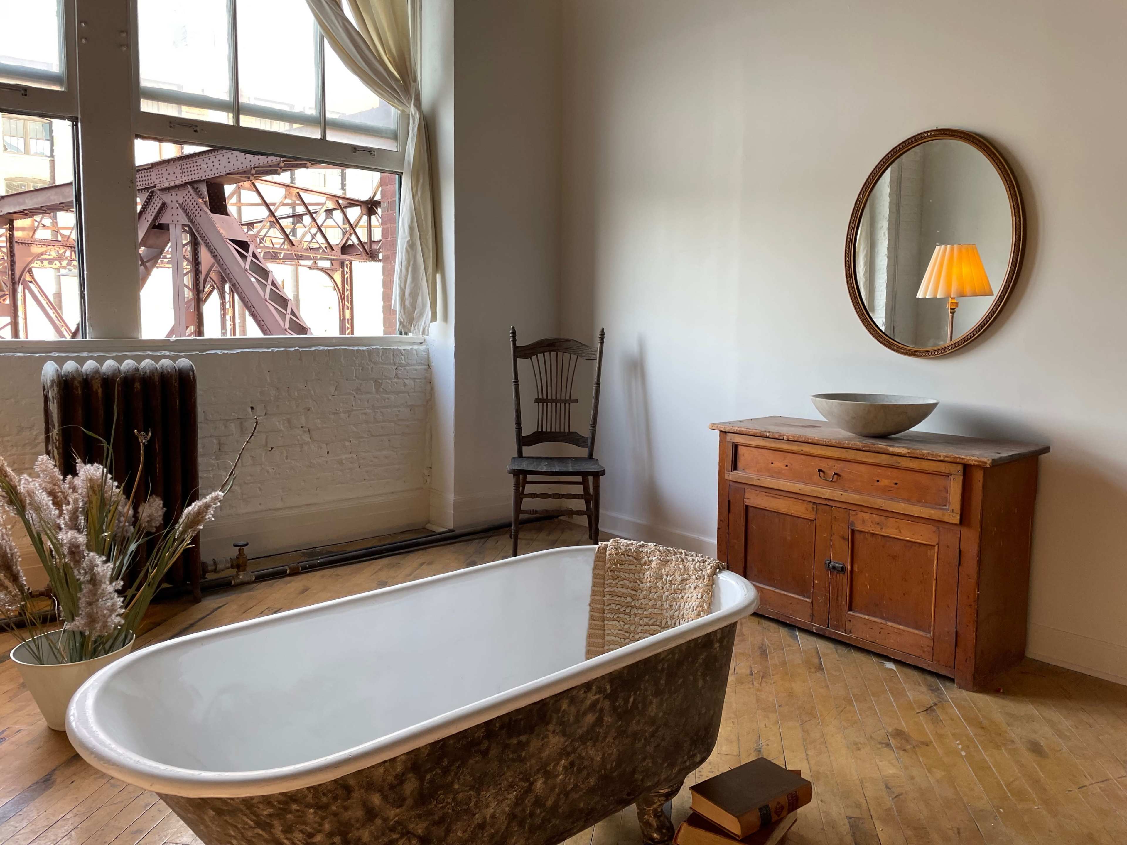 The image shows a vintage bathroom scene featuring a freestanding bathtub, a wooden chair, a round mirror, an antique cabinet, and a window with sheer curtains.