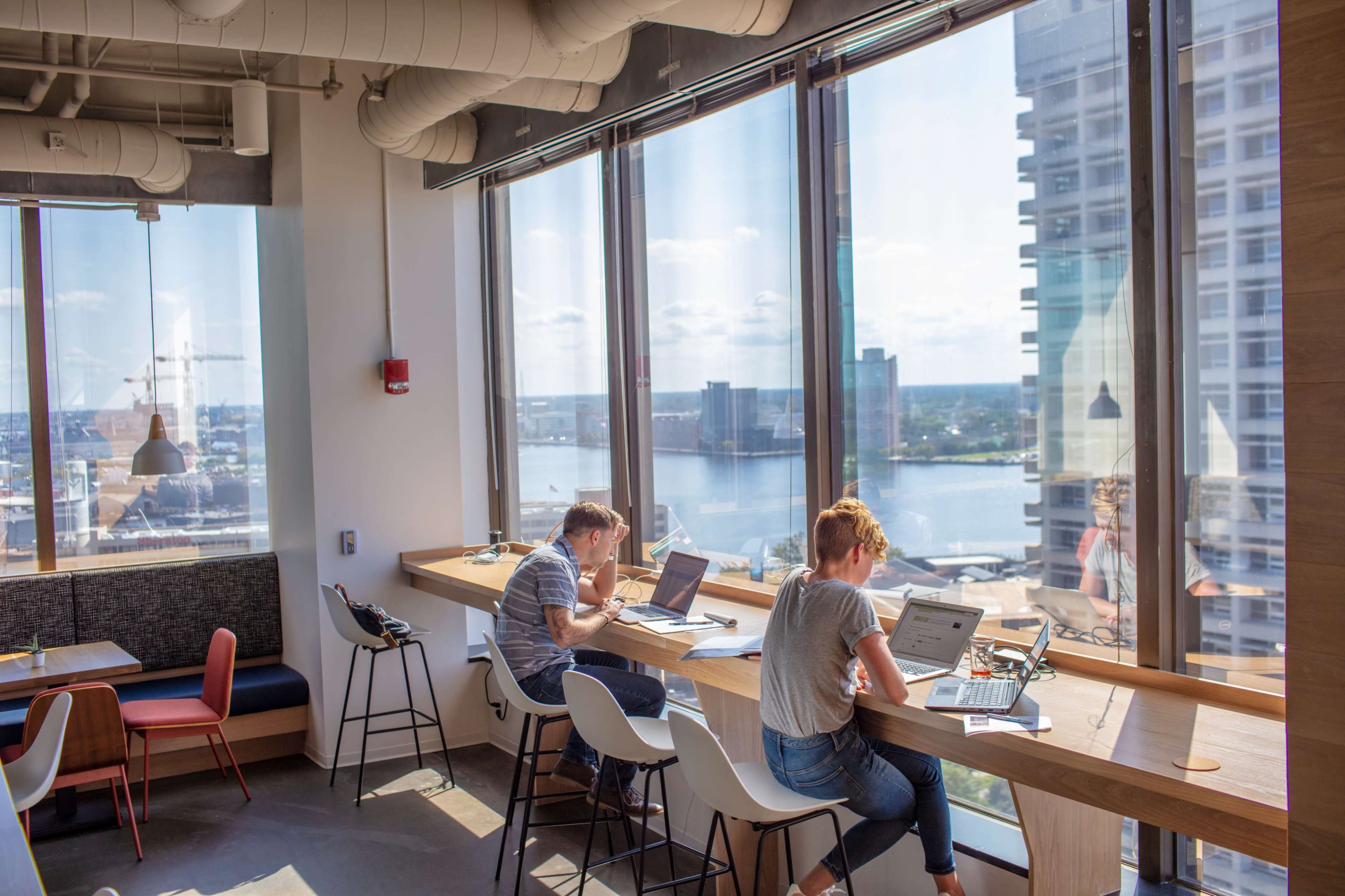 Two people work at a wooden counter near large windows overlooking a cityscape and water.