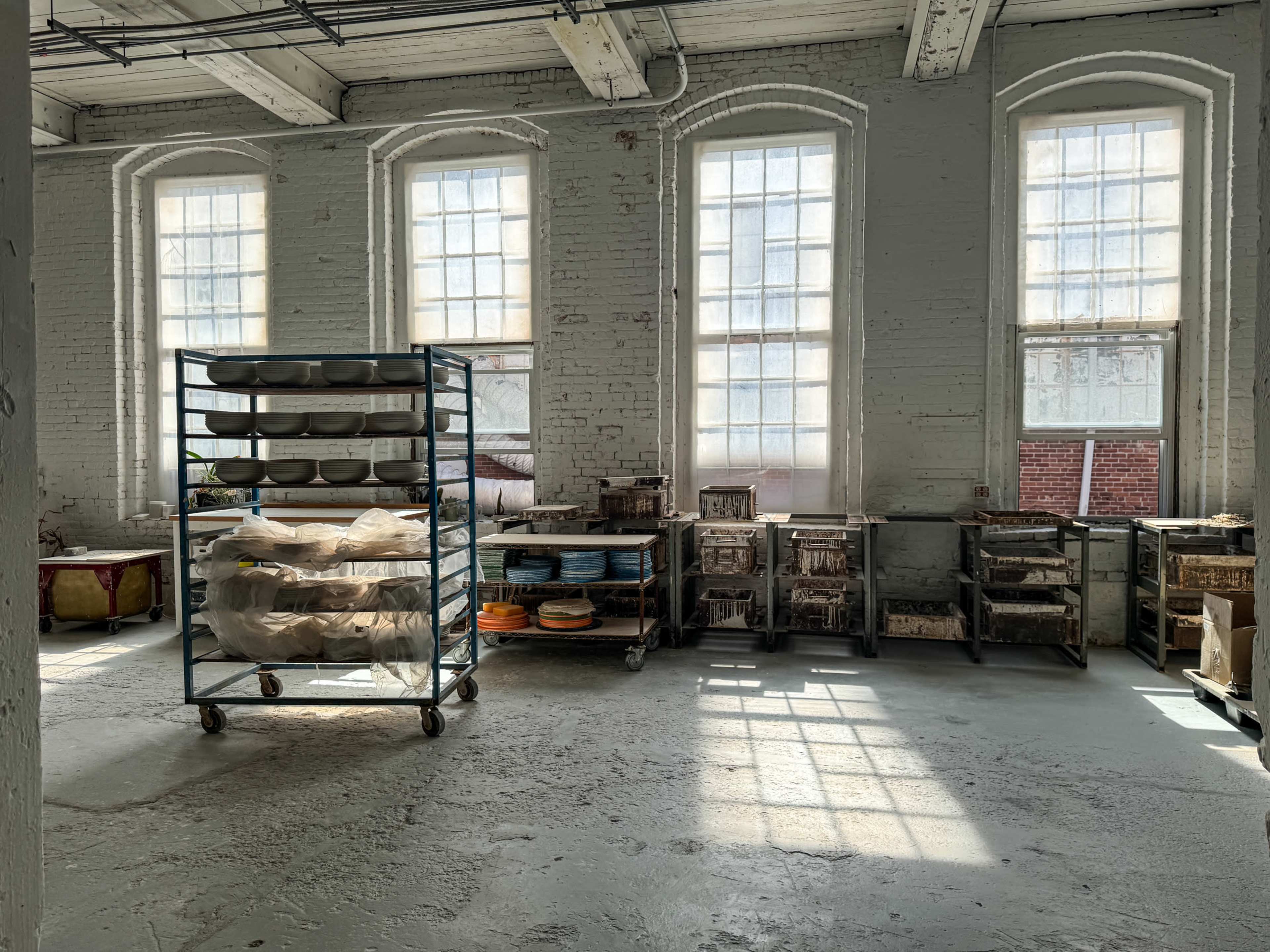A bright, sunlit room features tall windows and shelving units filled with pottery and tools.