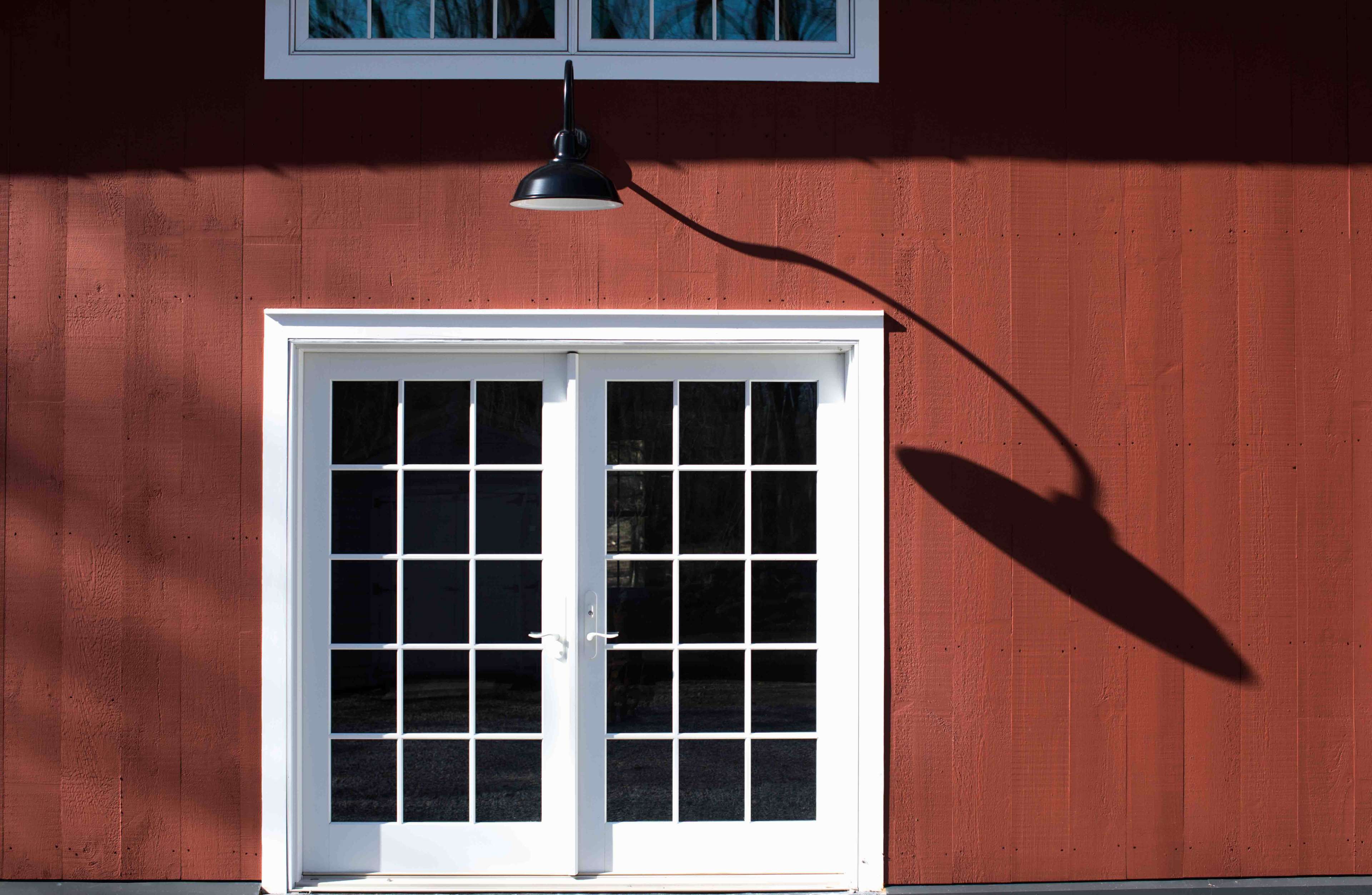 A set of double glass doors with a black light fixture above is mounted on a red wall, casting a distinct shadow.