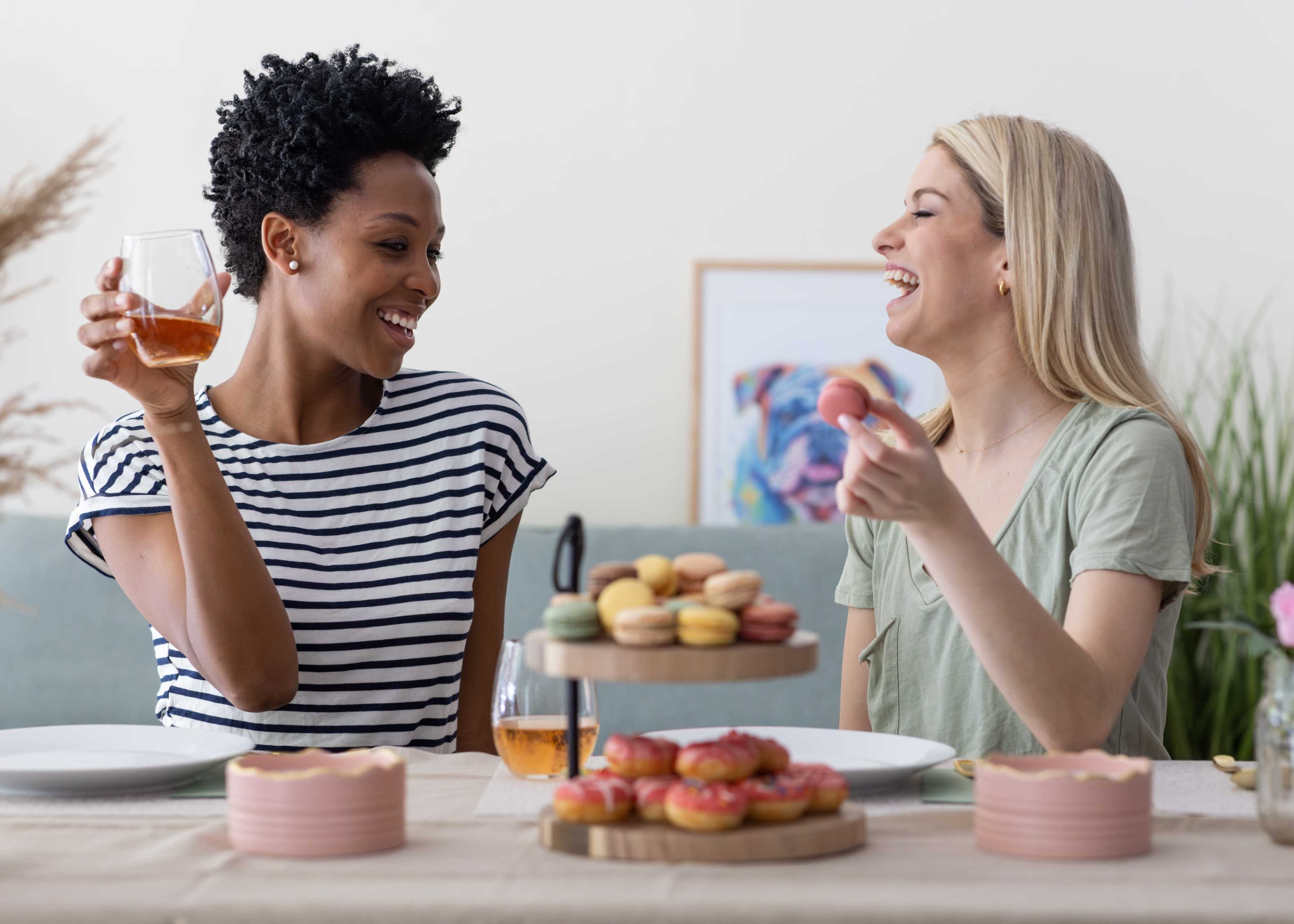 Two women sit at a table filled with desserts, smiling and enjoying drinks while engaged in conversation.