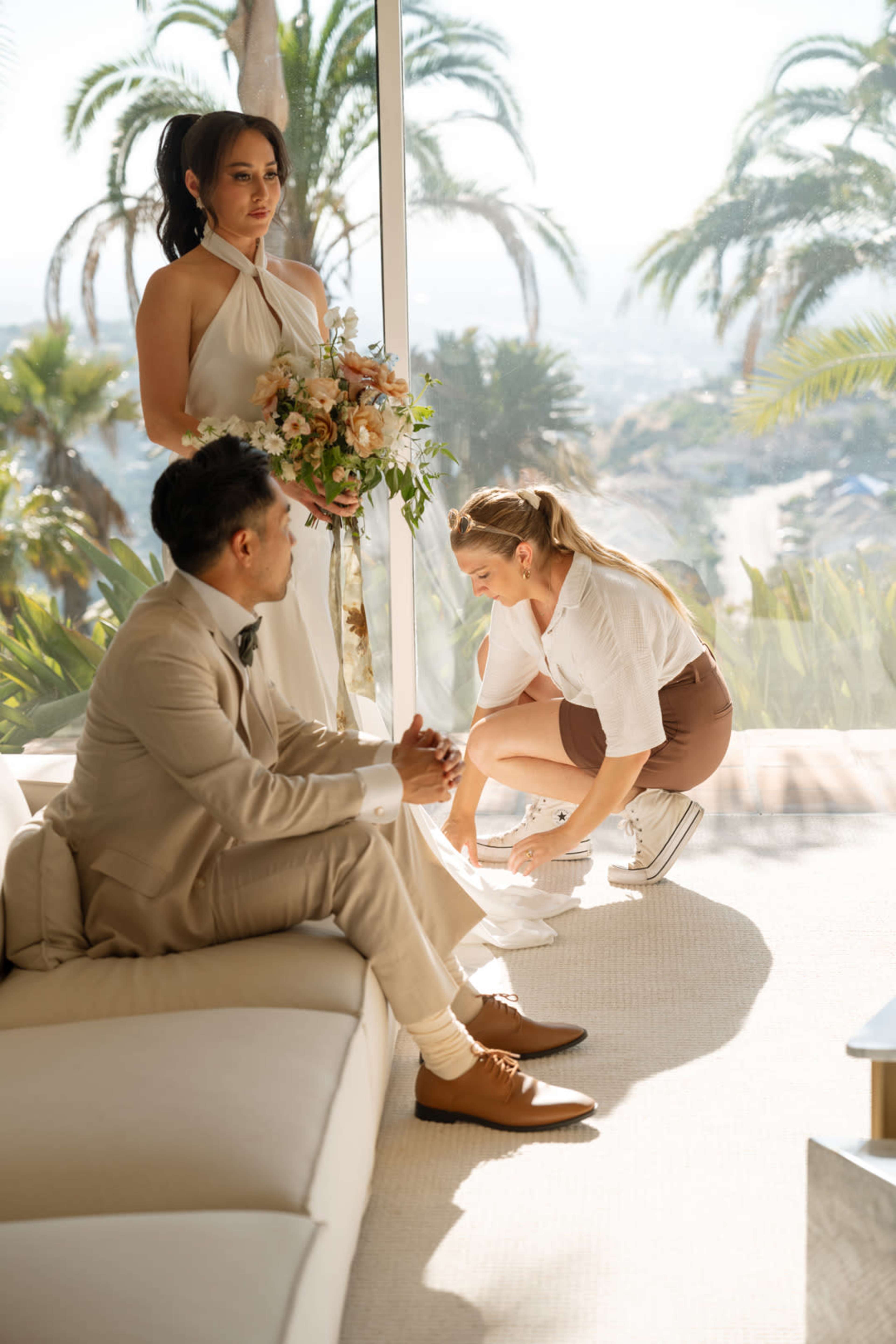 A bride holds a bouquet while a woman adjusts her dress, as a man sits on a sofa in a bright, tropical setting.