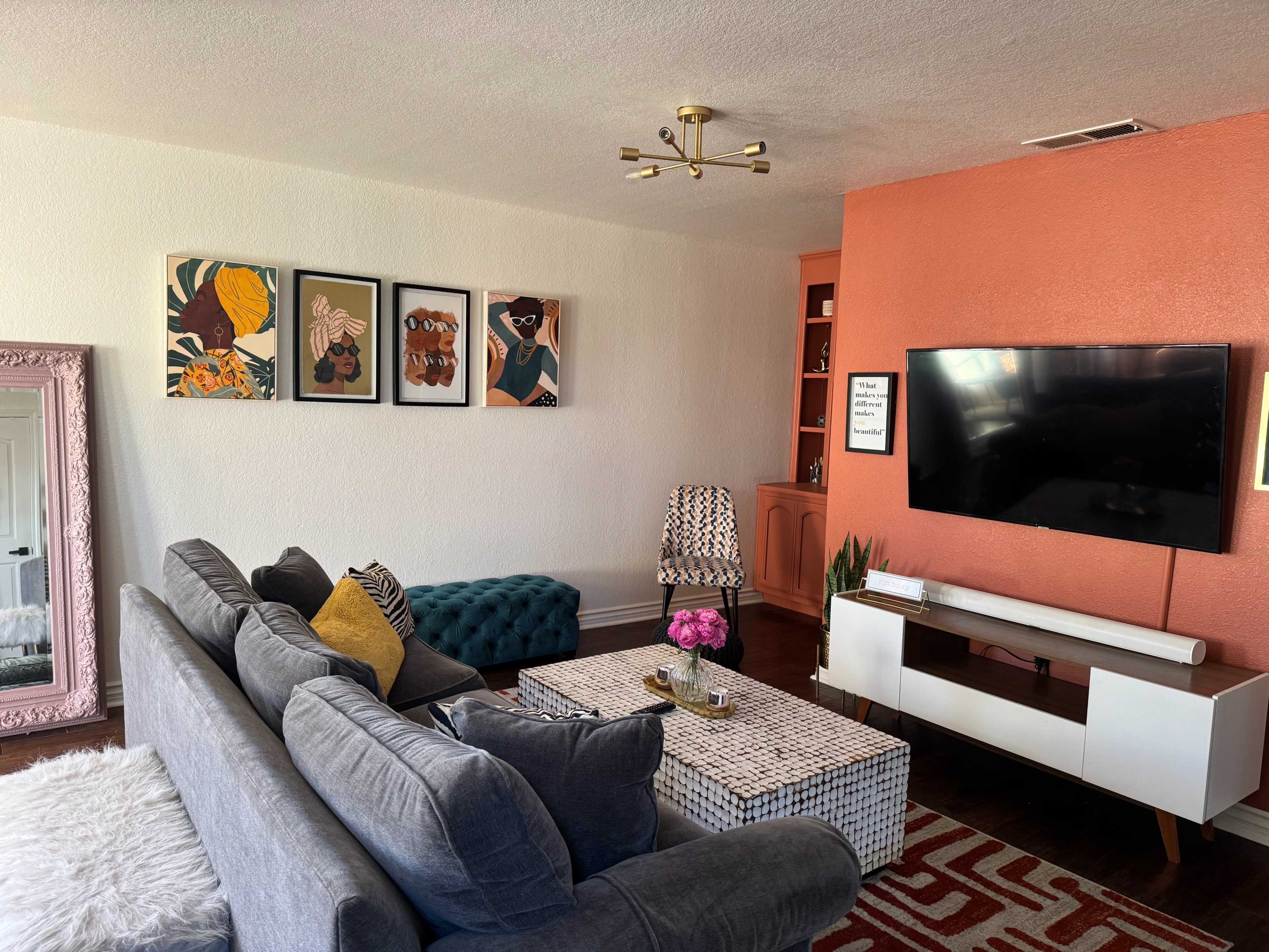 The living room features a gray couch with colorful pillows, a patterned coffee table, a television mounted on an orange accent wall, and framed art on the white wall above the couch.