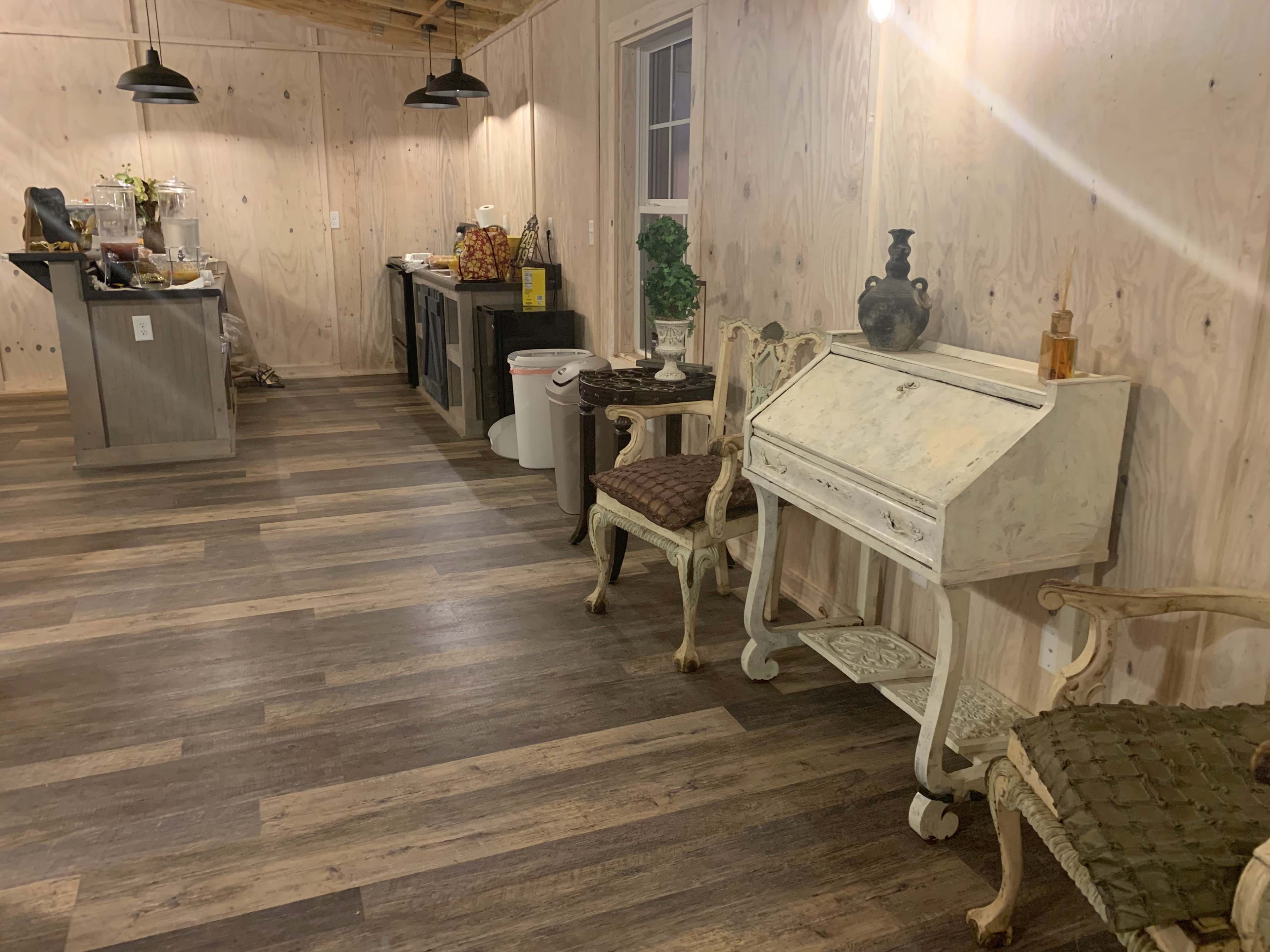 The image shows a wooden interior space with a polished floor, featuring a small desk, two vintage chairs, and a kitchenette area in the background.