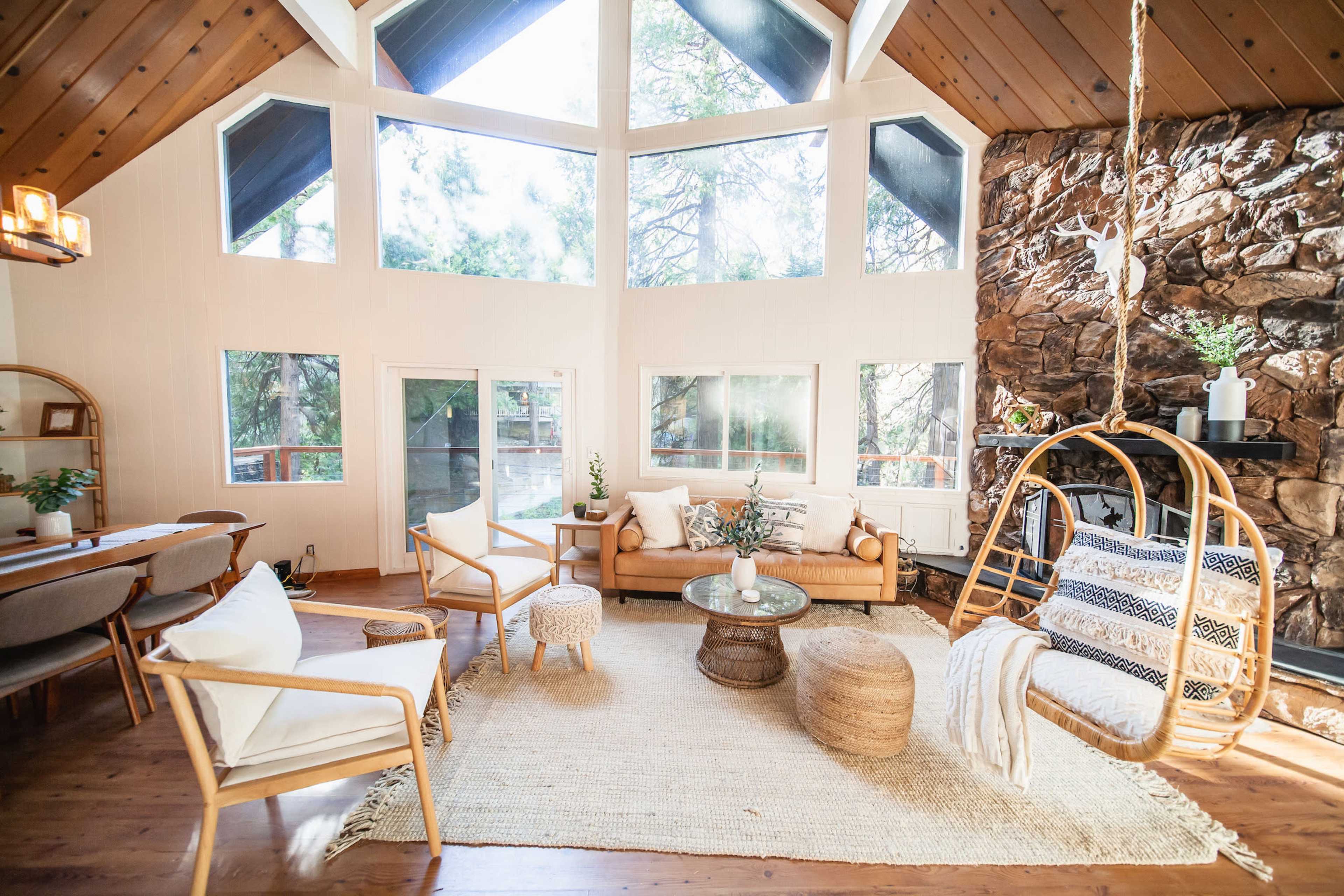 The image shows a modern living room with large windows, a stone fireplace, and a mix of comfortable seating arrangements including a hanging chair and a coffee table.