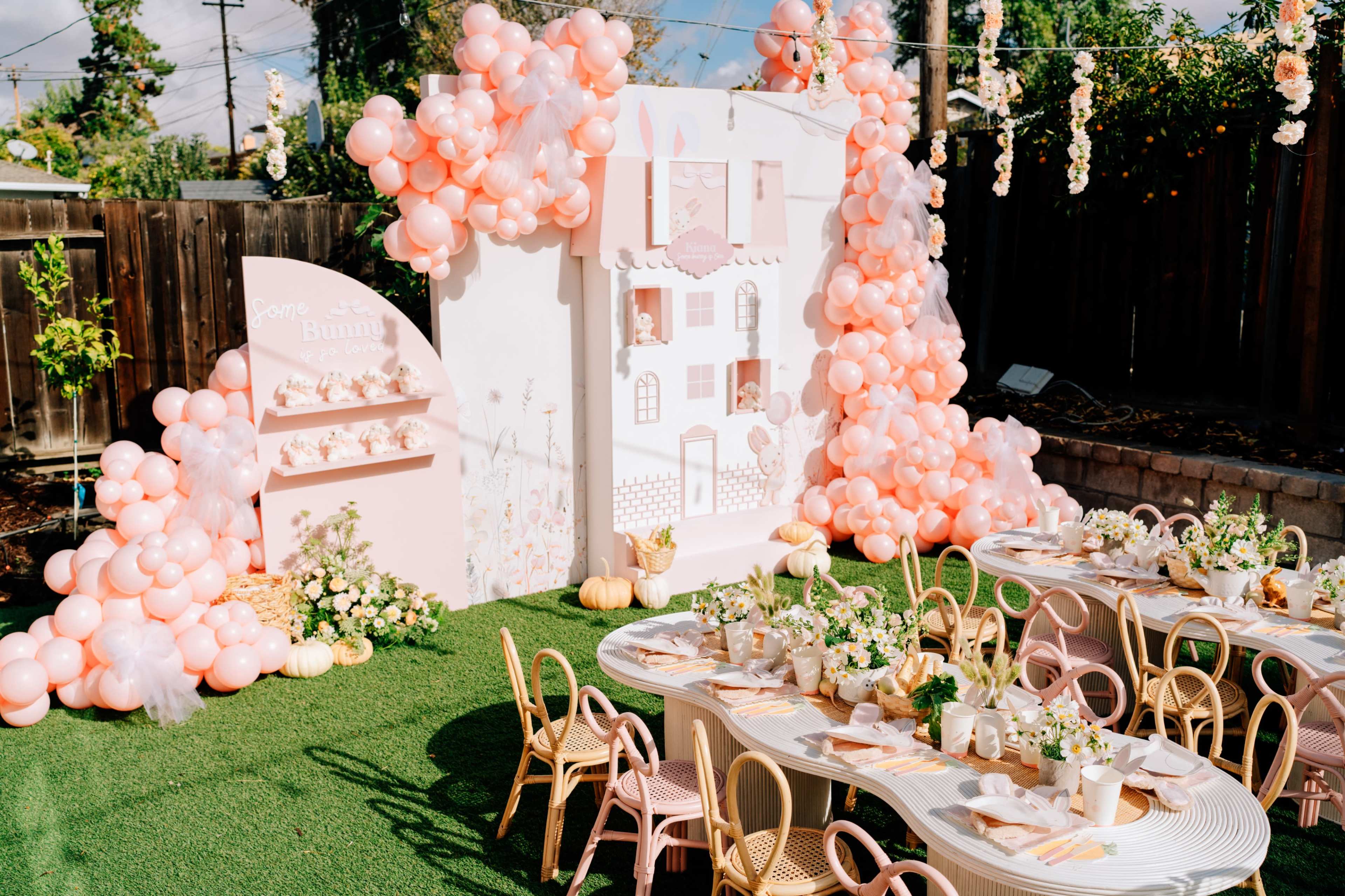 A backyard party setup features a pink-themed area with a decorative wall, pastel balloons, and a long table arranged with flowers and tableware.