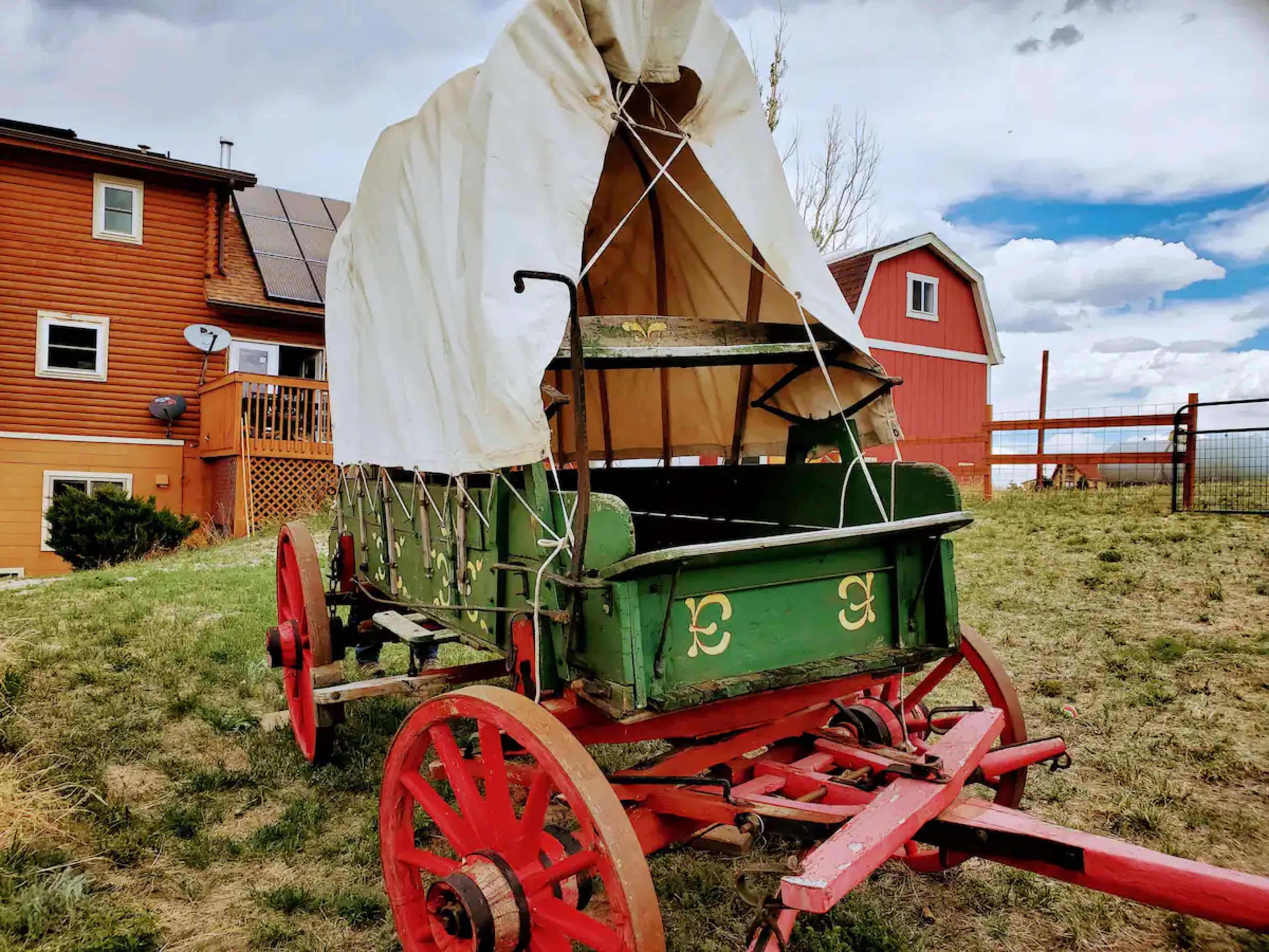 A covered wooden wagon with red wheels is positioned on grass near a house and barn in a rural setting.