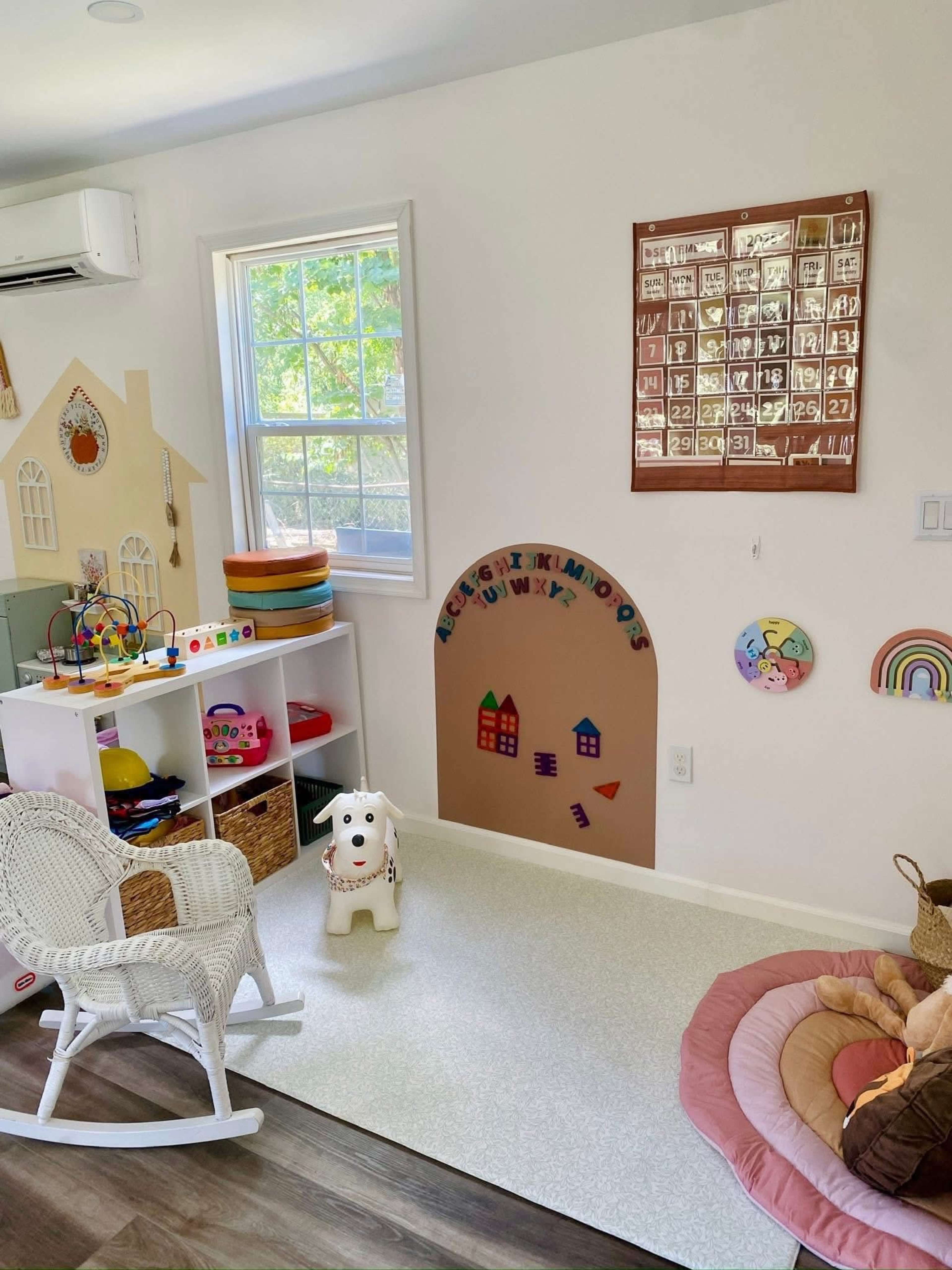 The room features a colorful play area with toys neatly organized in shelves, a calendar on the wall, and a cozy seating arrangement.