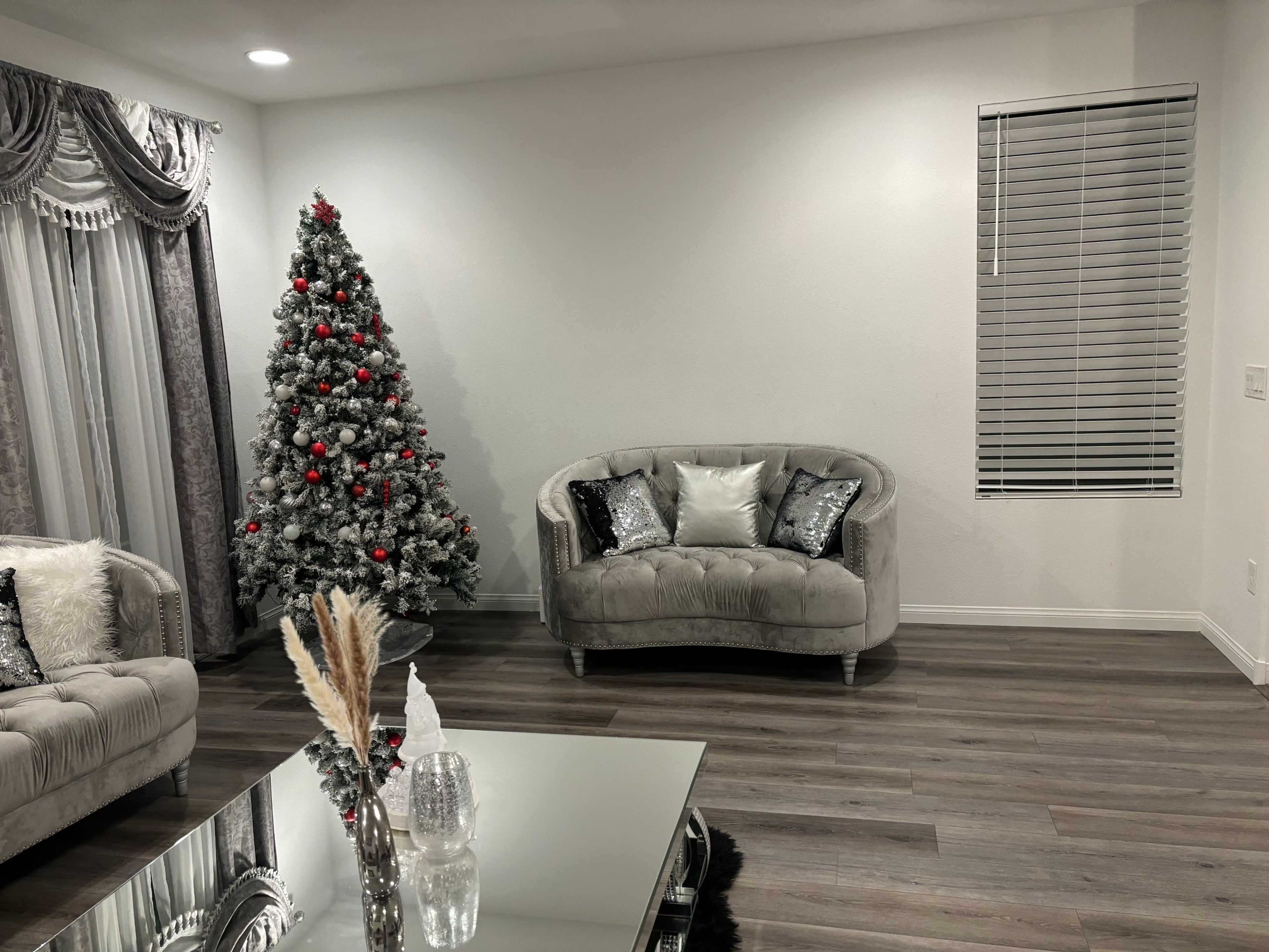 The scene features a decorated Christmas tree in a living room with a gray sofa, a glass coffee table, and a window with blinds.