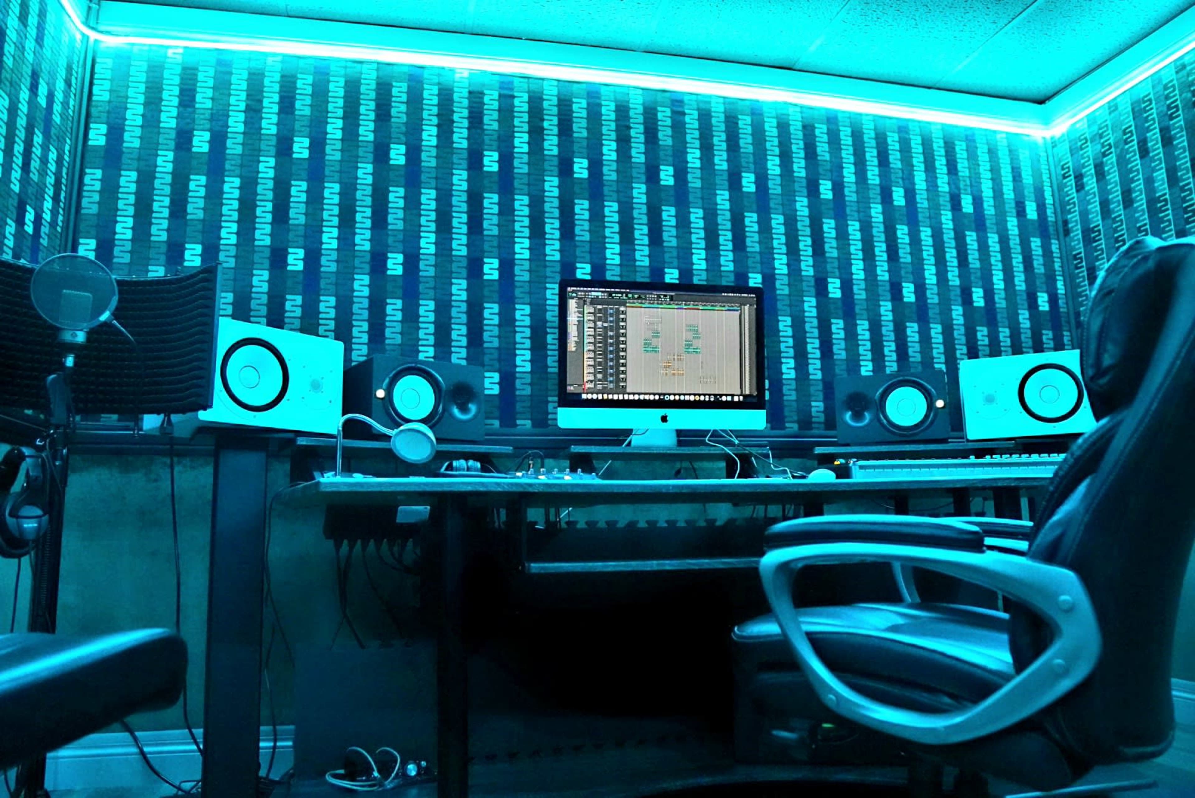 A music production studio features a computer with a digital audio workstation displayed, flanked by monitor speakers and illuminated by blue LED lighting.