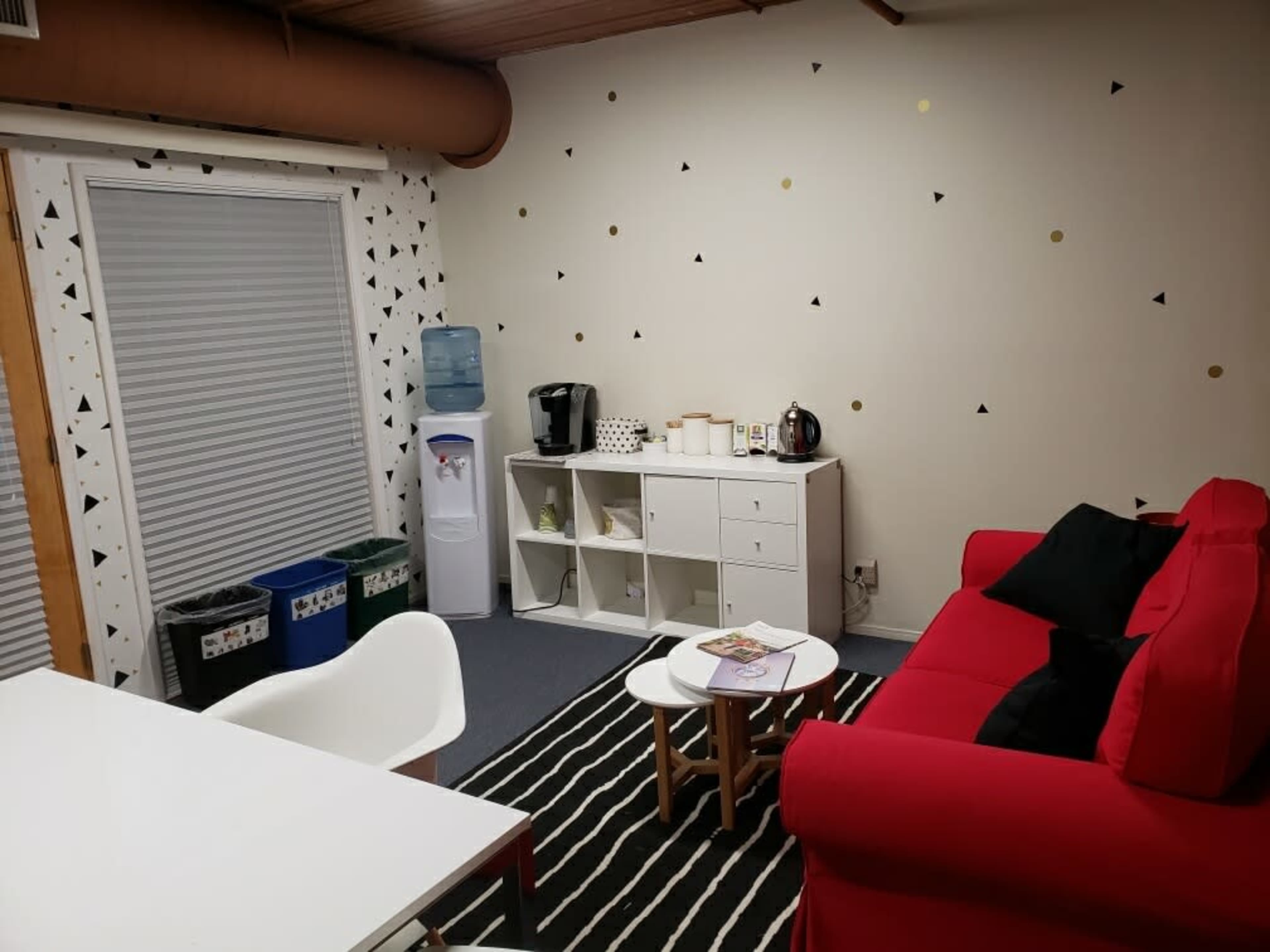 A small room with a red sofa, a white table, a water cooler, and a shelf with kitchen items, decorated with geometric wall designs.
