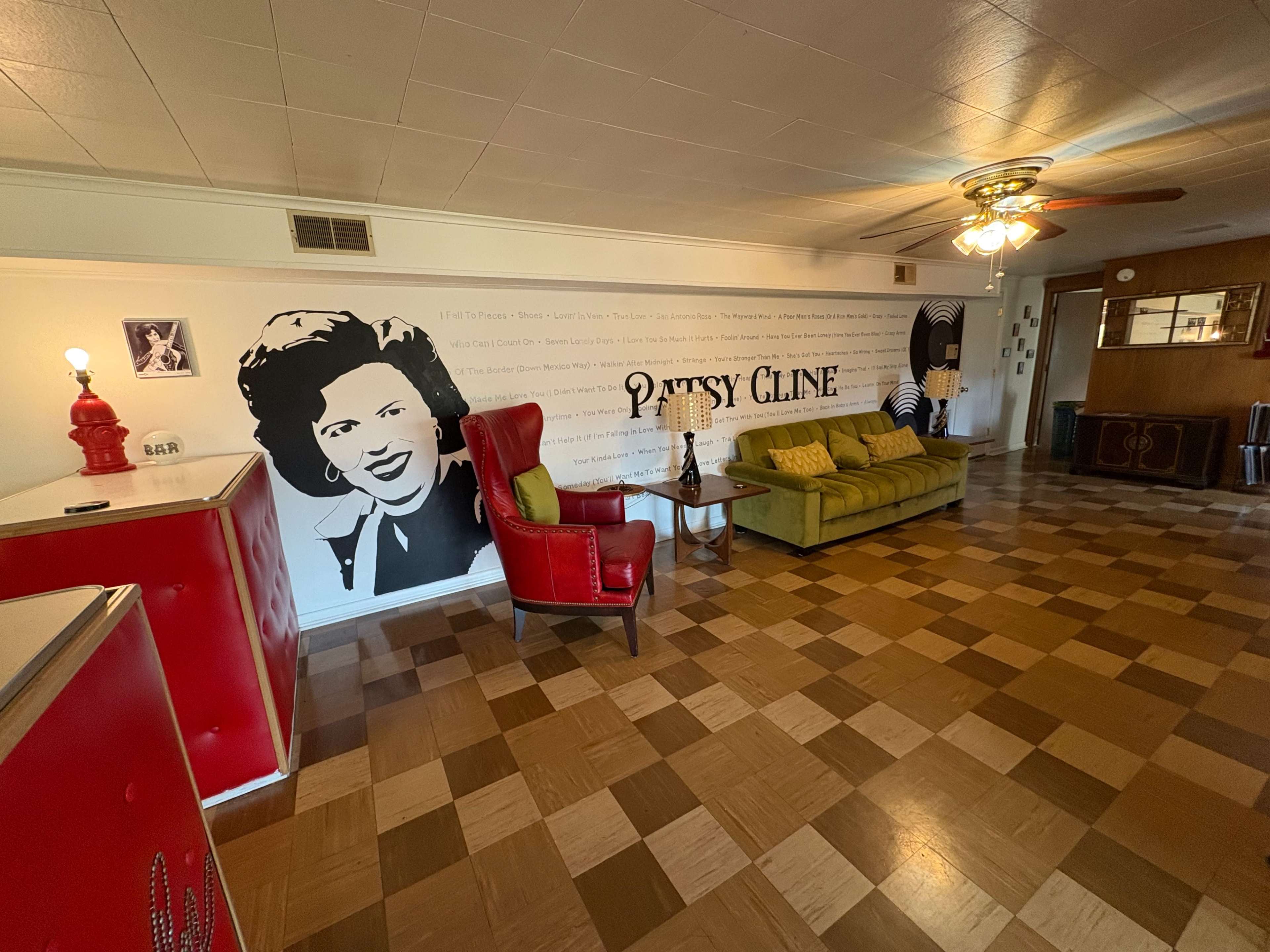 The image shows a living room with a distinctive wall mural of Patsy Cline, featuring a red chair, a green sofa, and a wooden coffee table on a patterned floor.