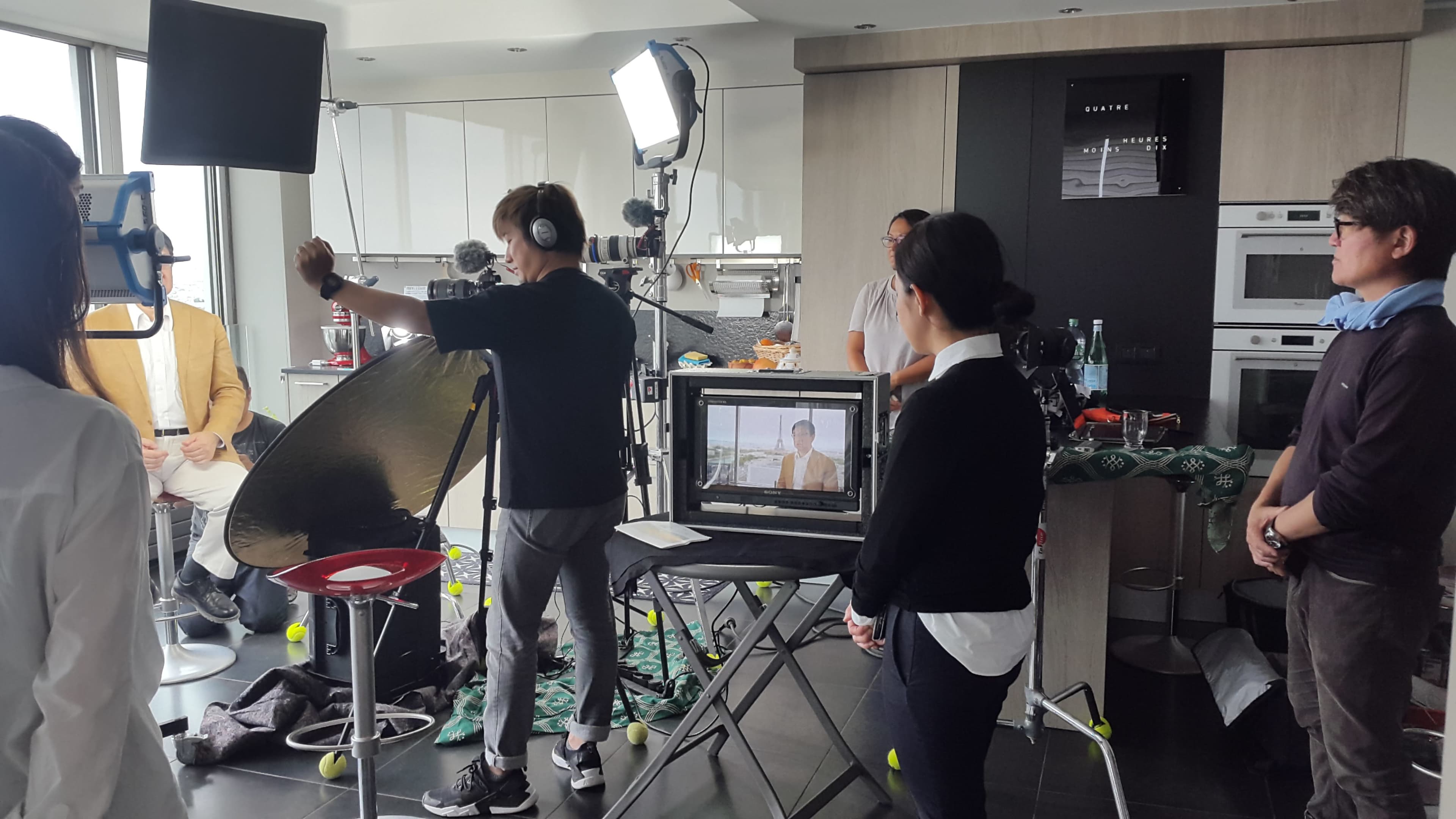 A film crew sets up a shoot in a modern kitchen, with a camera pointed at a seated subject and various lighting equipment positioned around.