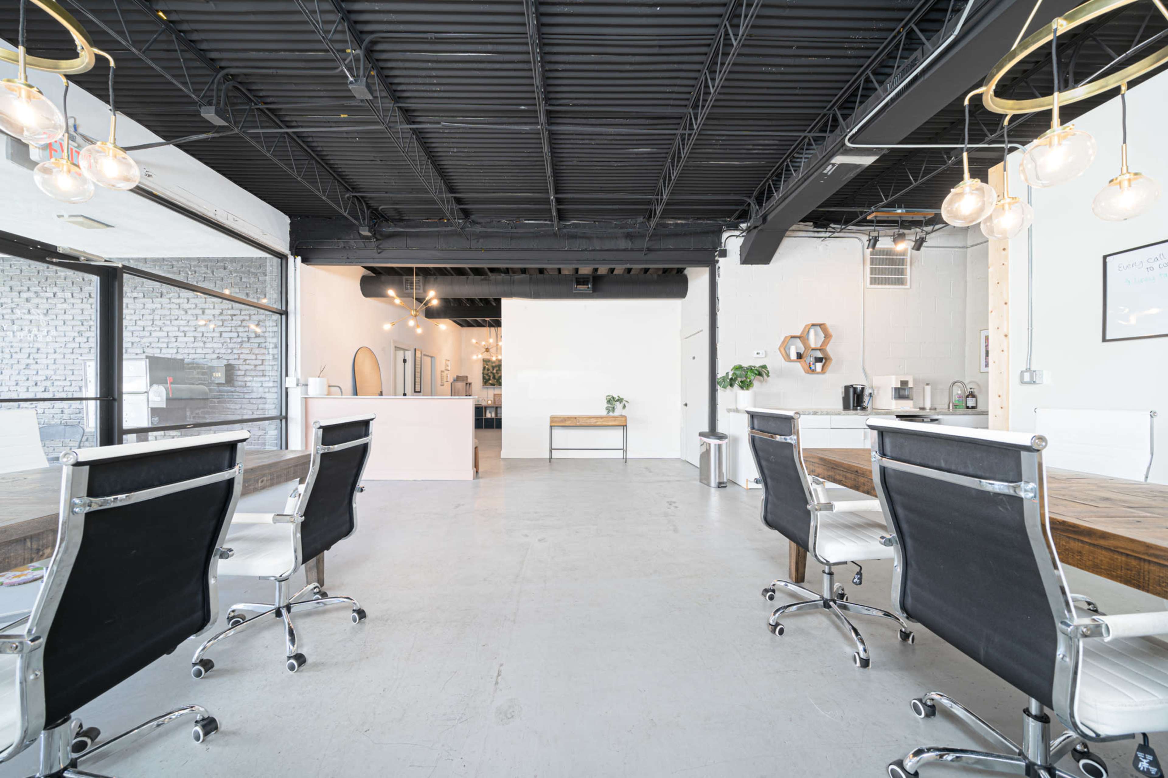 The image shows a modern, open office space with a long wooden table, white ergonomic chairs, and a bright, minimalist design featuring exposed black beams and pendant lighting.