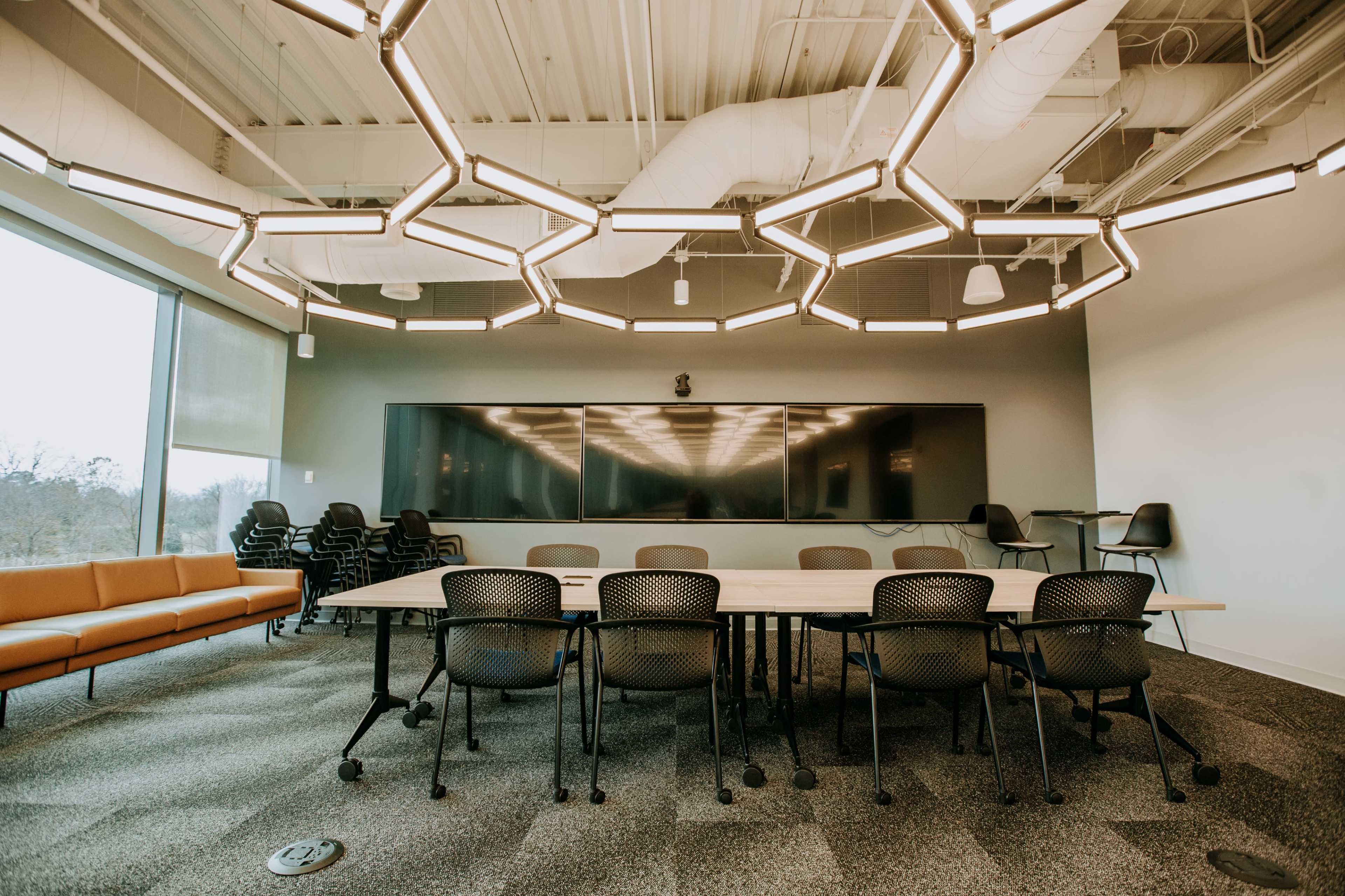 A modern conference room features a large table surrounded by black chairs, with a hexagonal light fixture hanging from the ceiling and a wall-mounted screen at the front.