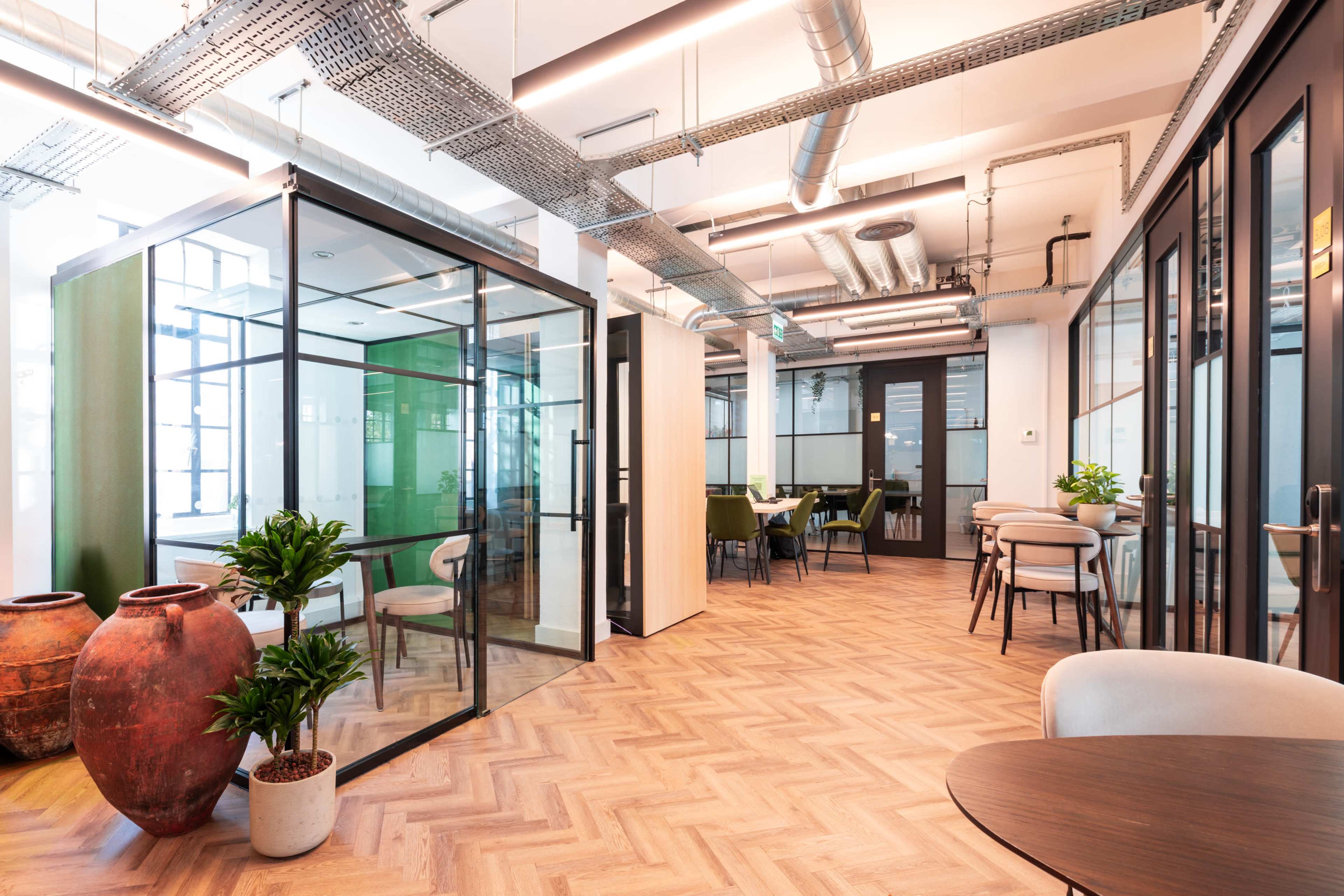 The image depicts a modern office space with glass-walled meeting rooms, wooden flooring, and potted plants throughout.