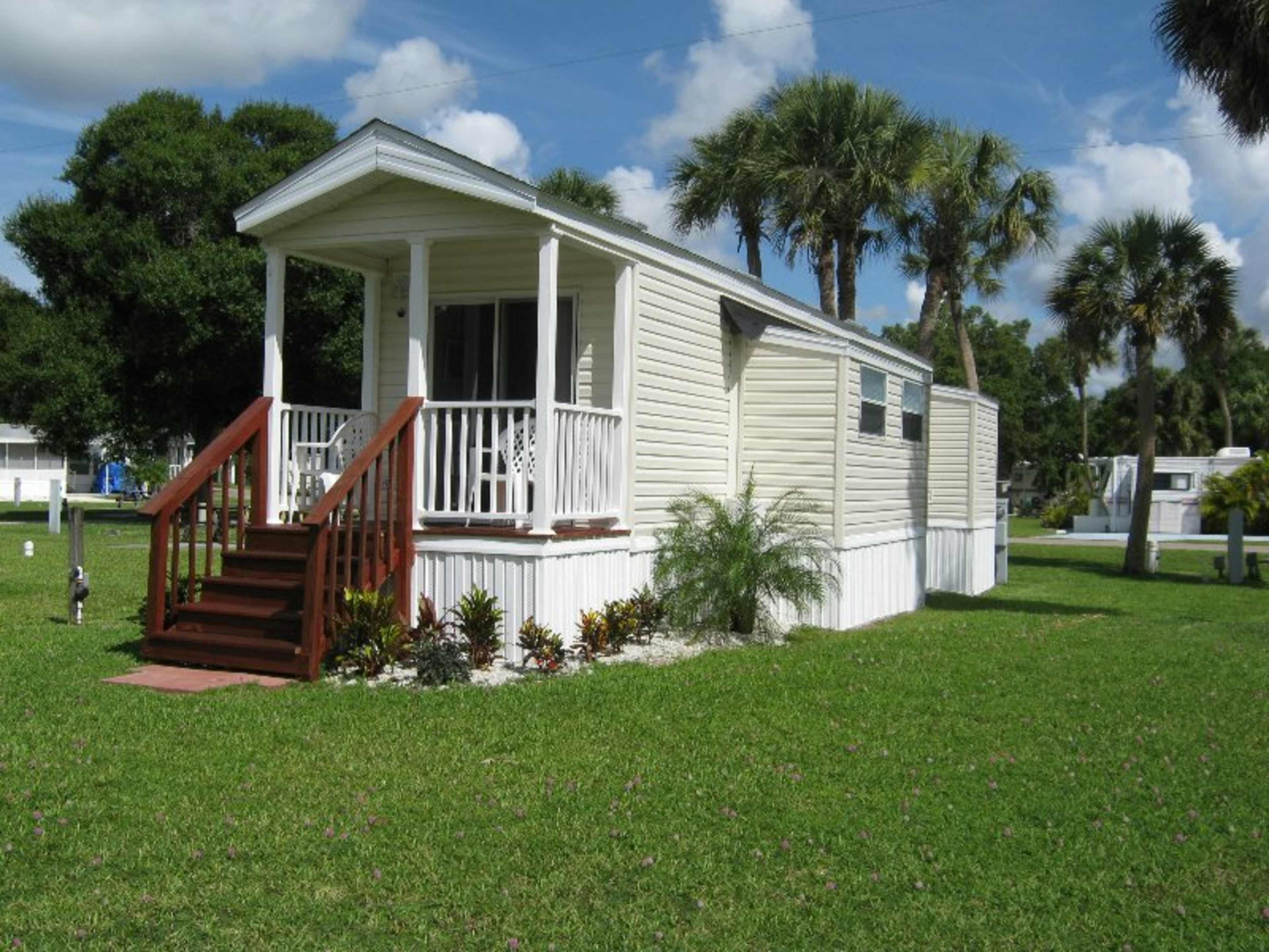 A small white manufactured home with a front porch and wooden steps is set on a grassy lawn surrounded by palm trees.